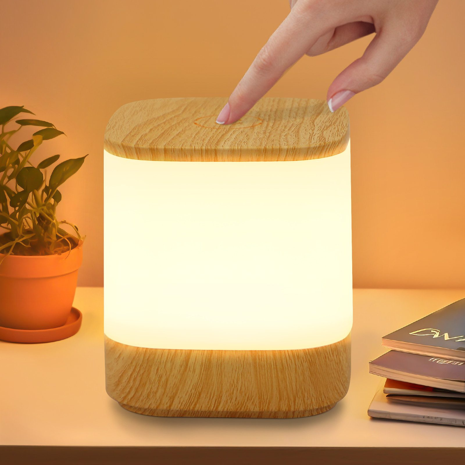 Drewniana lampa stoi na białym stole. Ręka dotyka wierzchu lampy. Jest też roślina i książki.