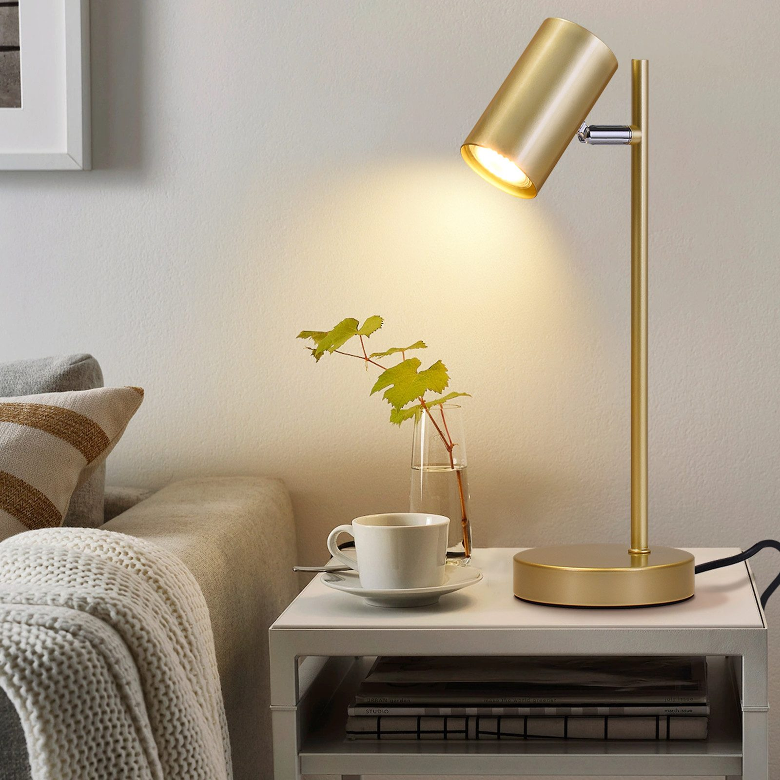 Nowoczesna lampa biurkowa w kolorze złotym ze włączonym światłem, świeci na białym stoliku nocnym.