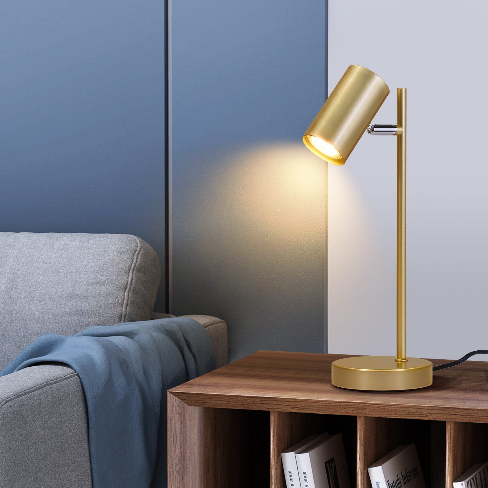 Nowoczesna lampa biurkowa w kolorze złotym. Światło pada na ciemny drewniany stół z książkami i wazonem z rośliną.