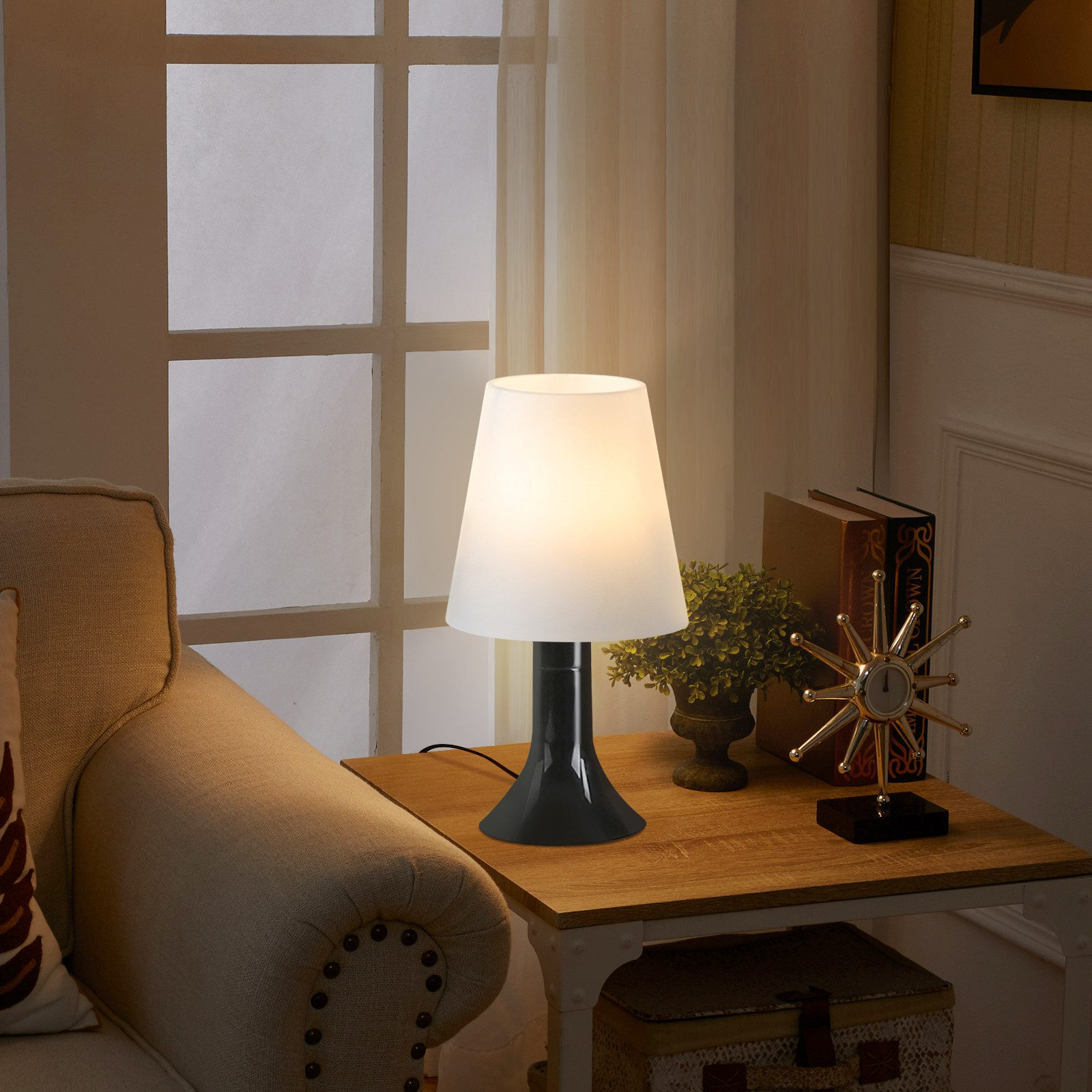 Biały klosz na czarnej podstawie stoi na drewnianym stole obok sofy.