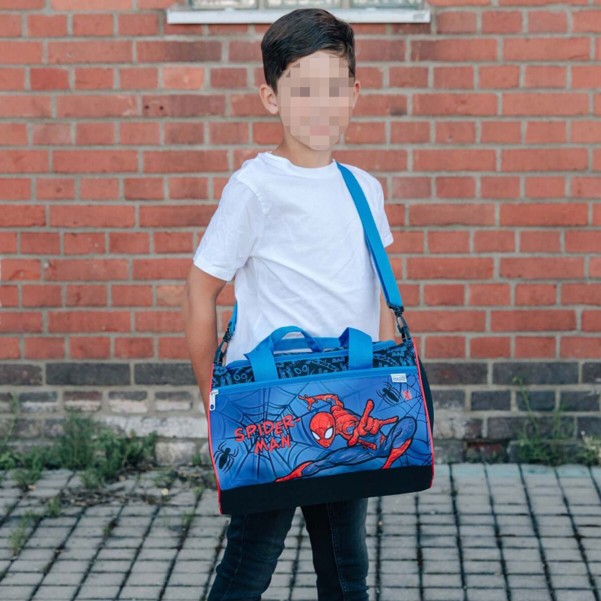 Ein Junge in weißem Hemd und Jeans steht mit einer Spiderman-Tasche vor einer Backsteinmauer.