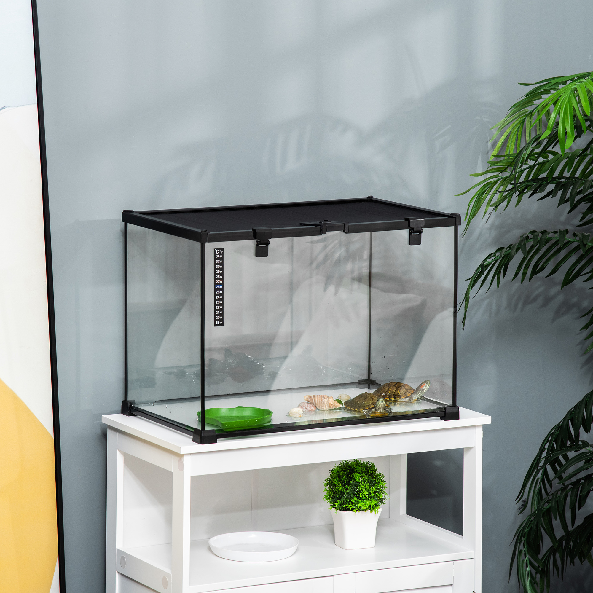 Ein Glasterrarium mit schwarzem Deckel und einer Schildkröte darin. Es steht auf einem weißen Tisch, mit Grün drumherum.
