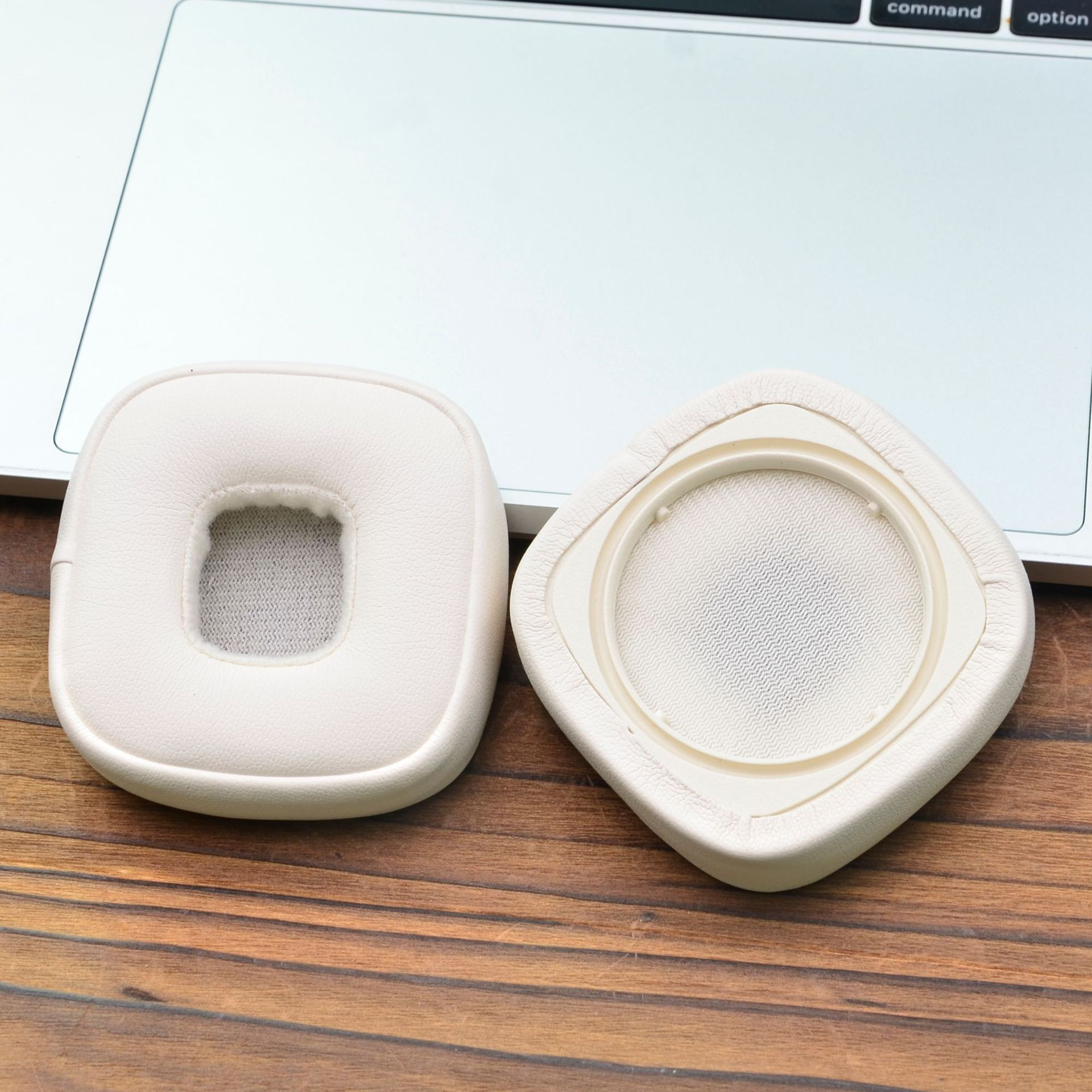 Due cuscinetti auricolari bianchi per cuffie sono su una superficie di legno, accanto a un laptop argentato.