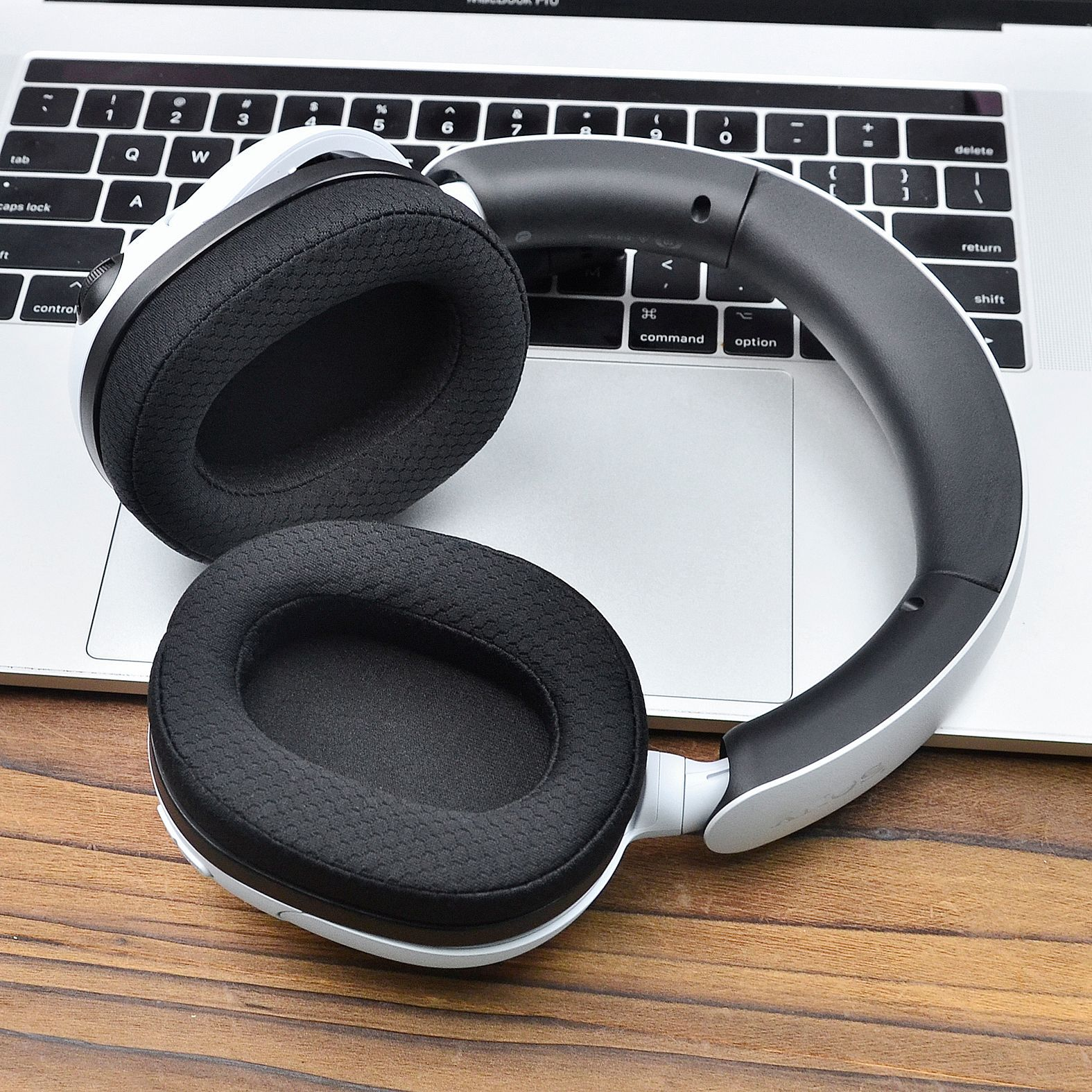 Słuchawki na klawiaturze laptopa. Czarne nauszniki i pałąk, białe akcenty. Drewniany stół.