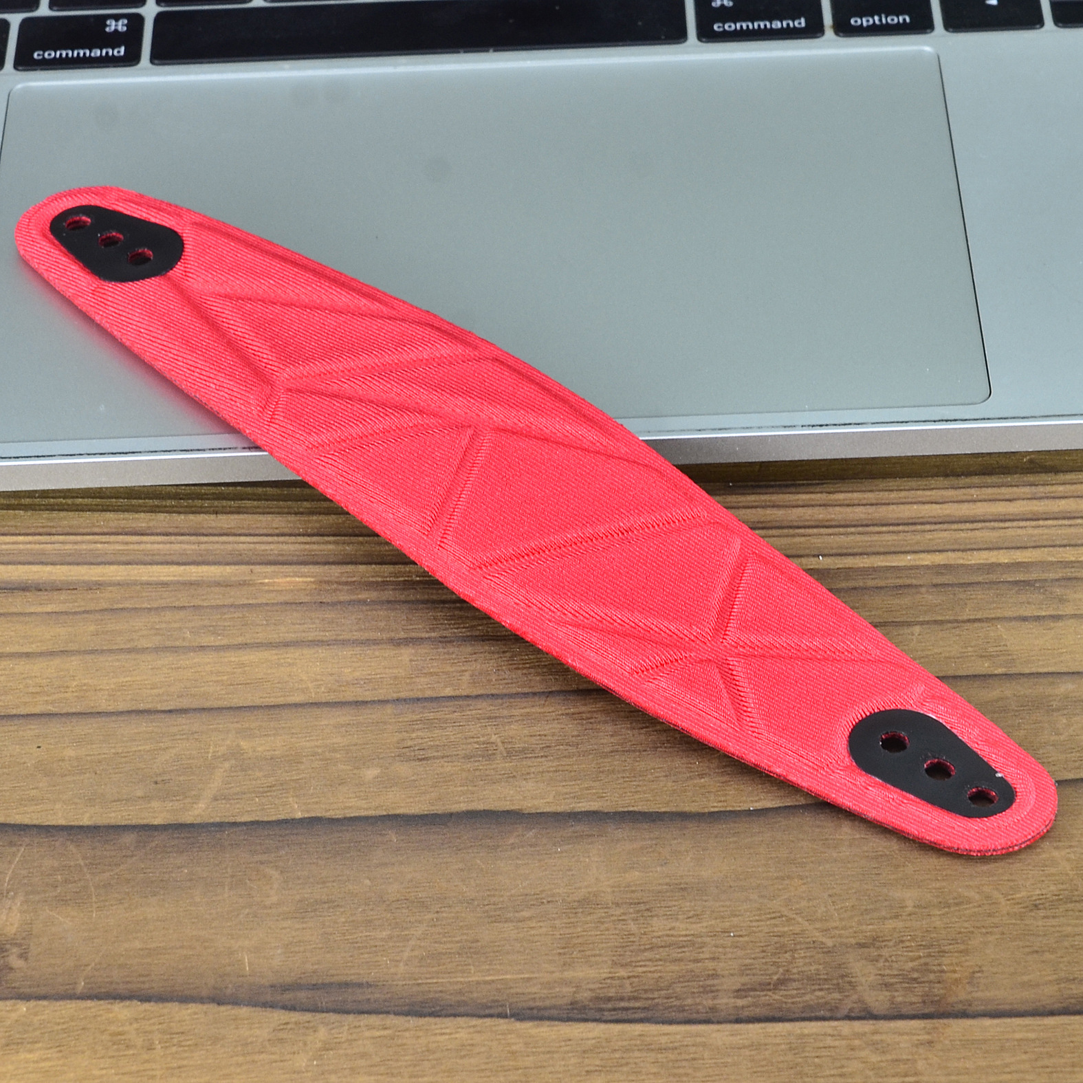 Un pezzo di plastica rosso e nero con fori, su una scrivania di legno accanto a un laptop.