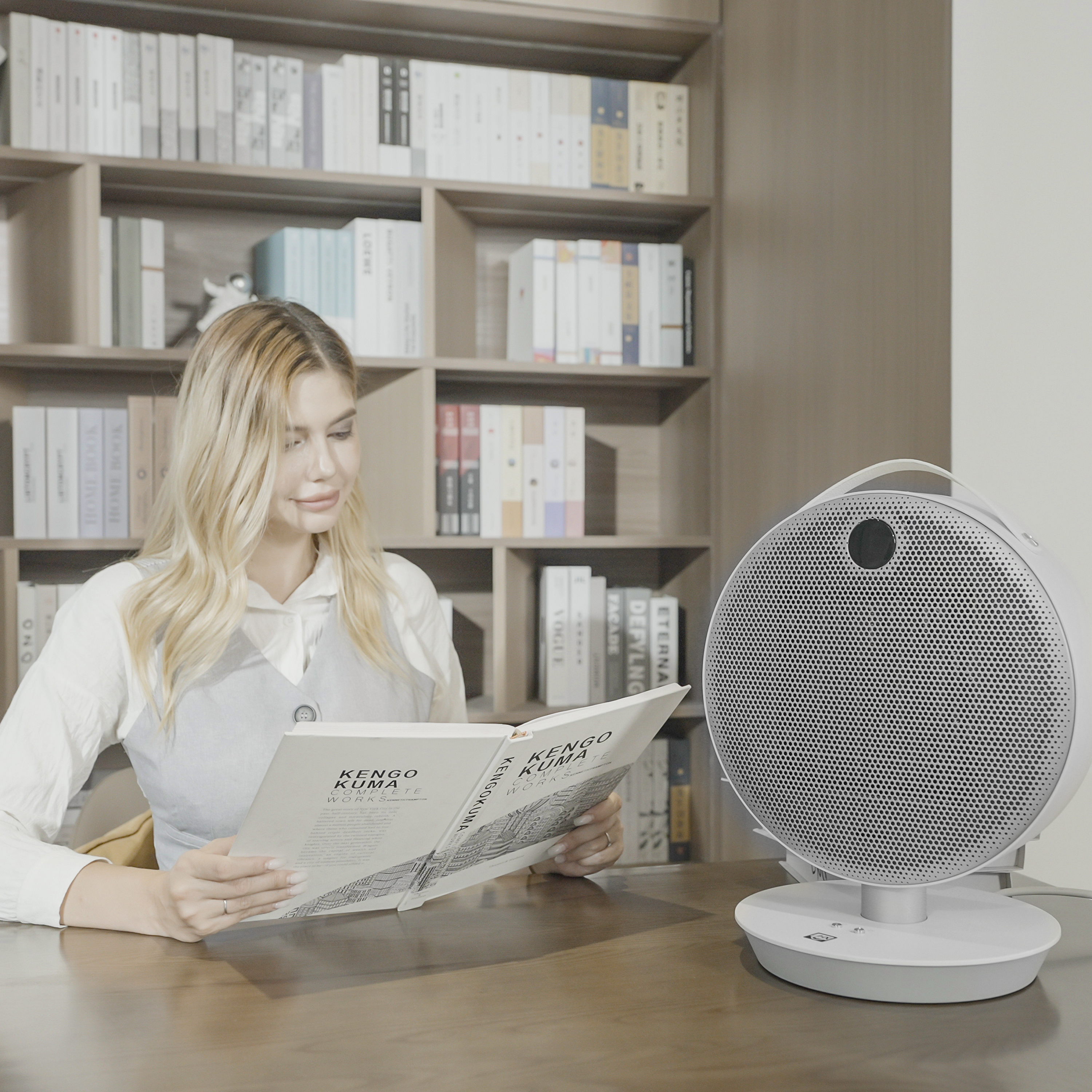 Frau liest Buch neben weißem Ventilator. Der Ventilator steht auf einem Sockel, perforierte Front.