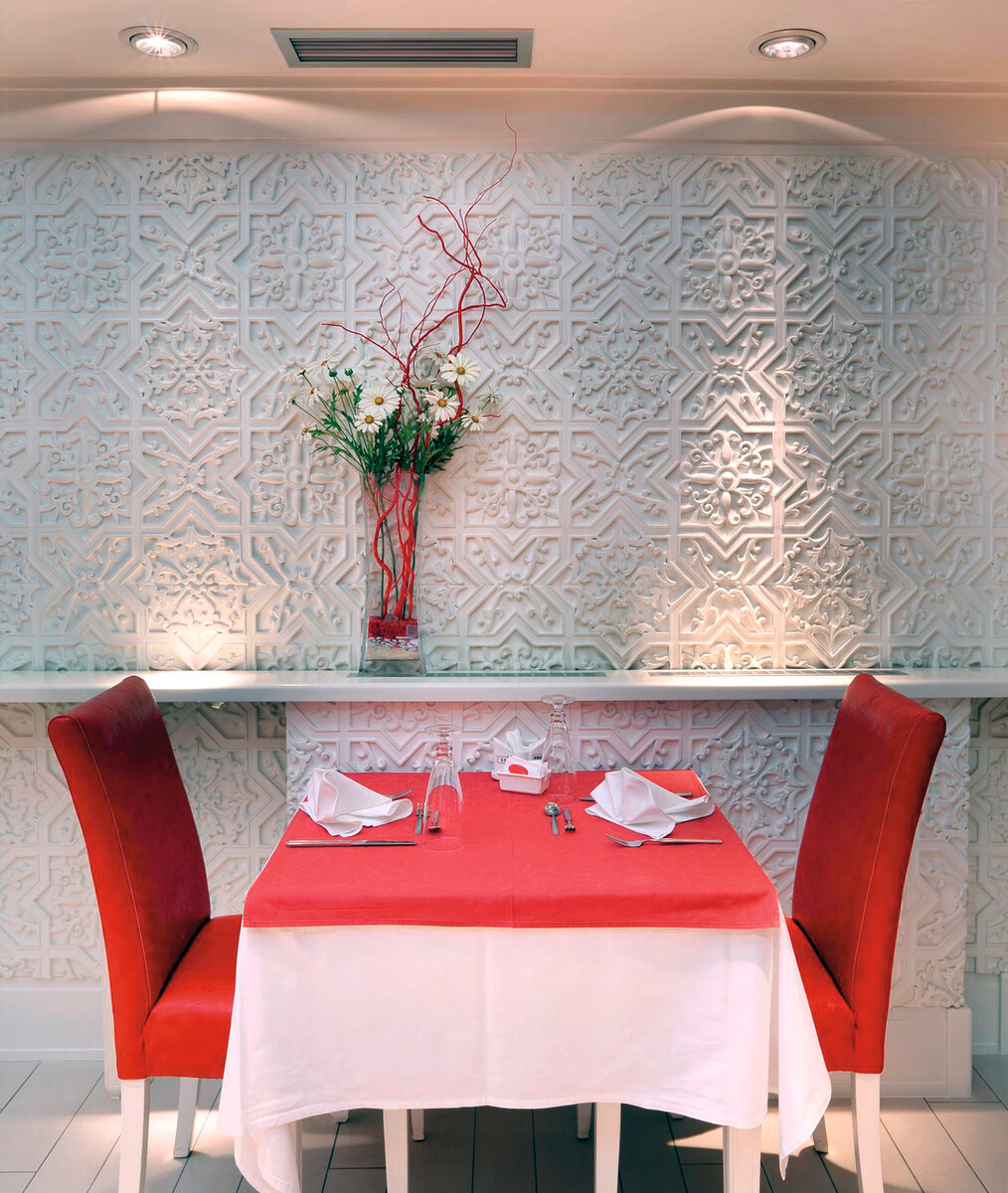 Tavolo da ristorante per due con sedie rosse, tovaglia bianca e parete bianca decorativa.
