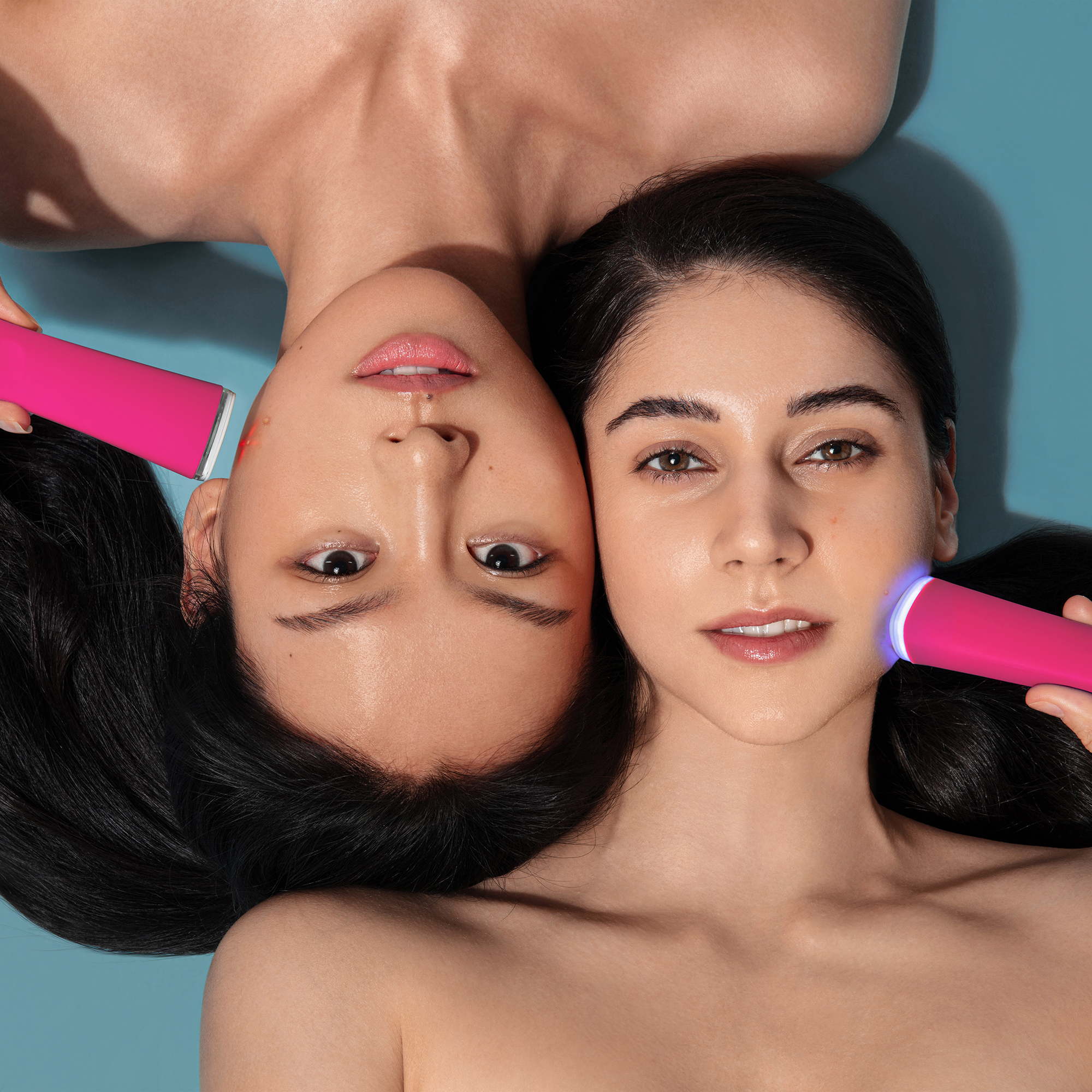 Two women are lying head to head using pink facial devices on their faces, blue background.