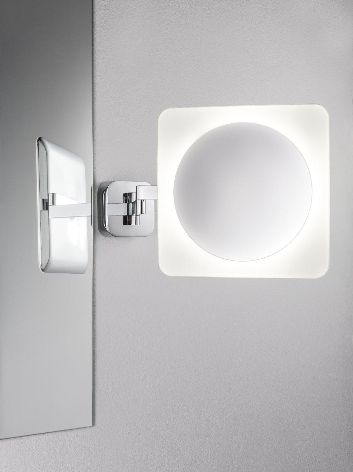 Un miroir mural moderne, de forme carrée, avec un miroir circulaire éclairé. Bras et cadre chromés.