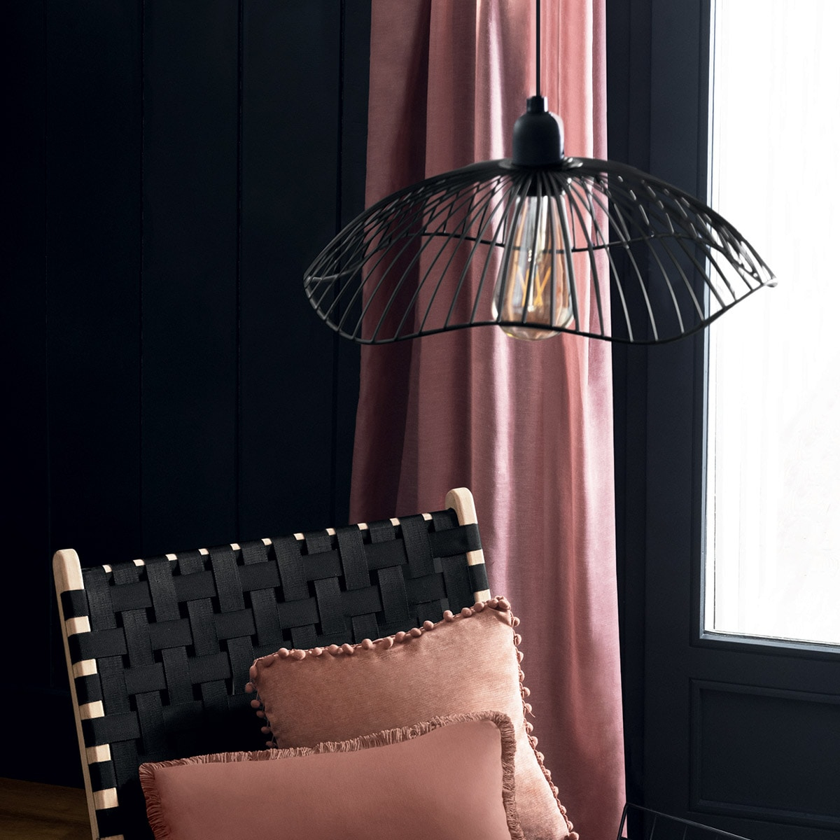 Black wireframe lamp with lit bulb, above a black chair and pink pillows.