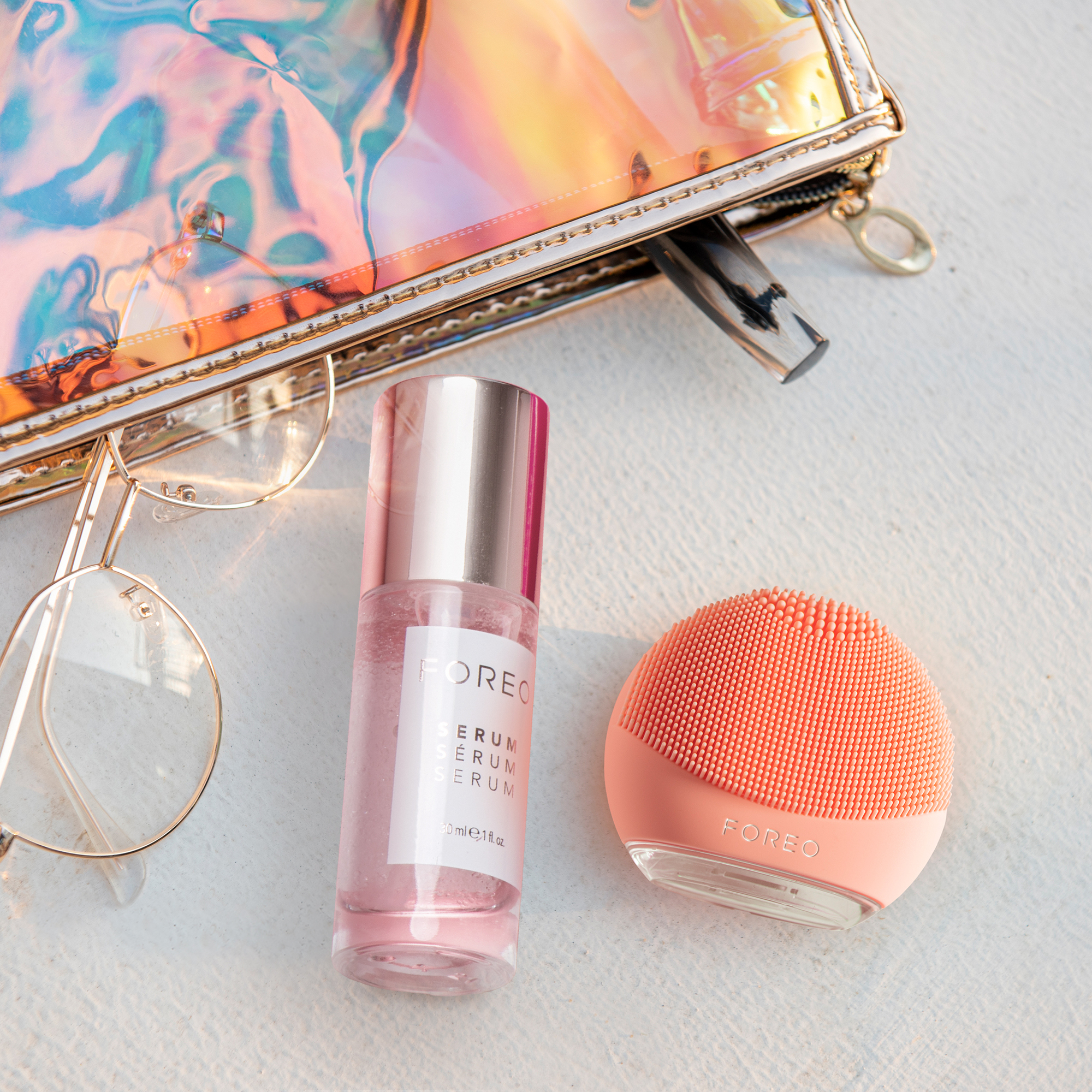 Foreo serum, glasses, and cleansing device on white surface, with a pouch.