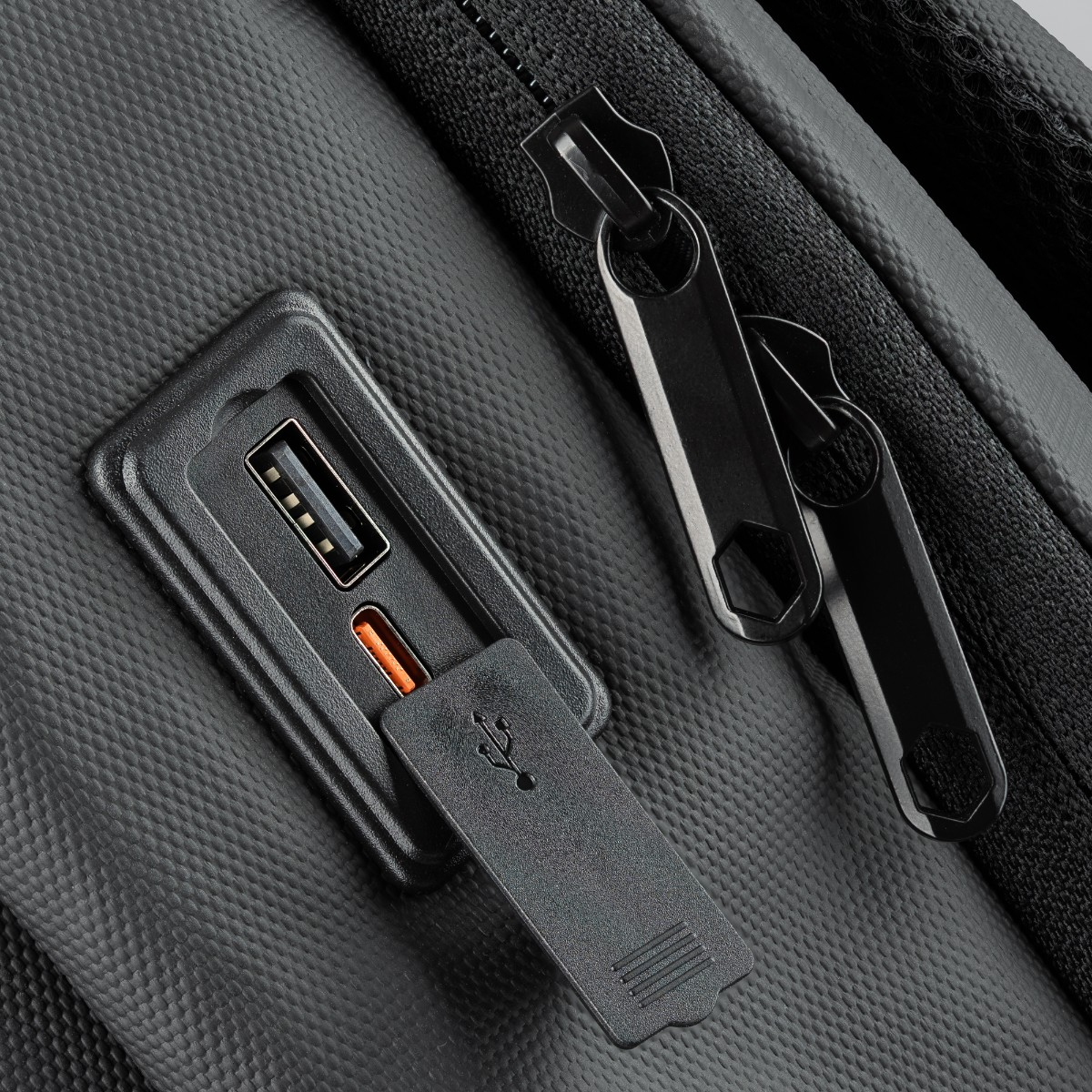 Zbliżenie portu USB czarnego plecaka, z USB i portem ładowania, na teksturowanej powierzchni.