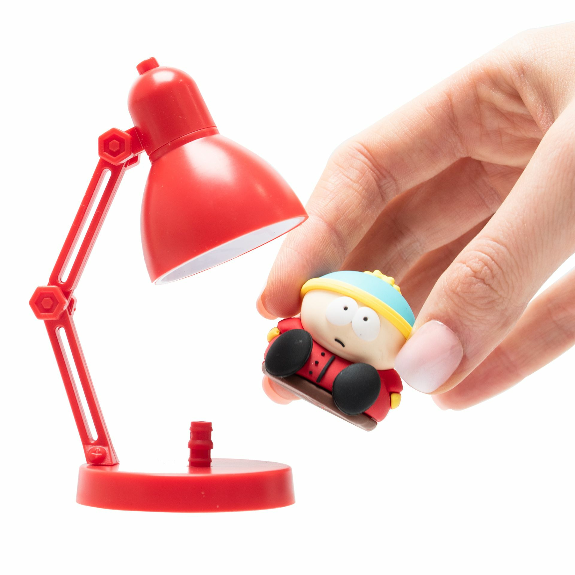 Una mano sosteniendo una figura de Cartman, junto a una lámpara de escritorio roja. La lámpara está encendida.