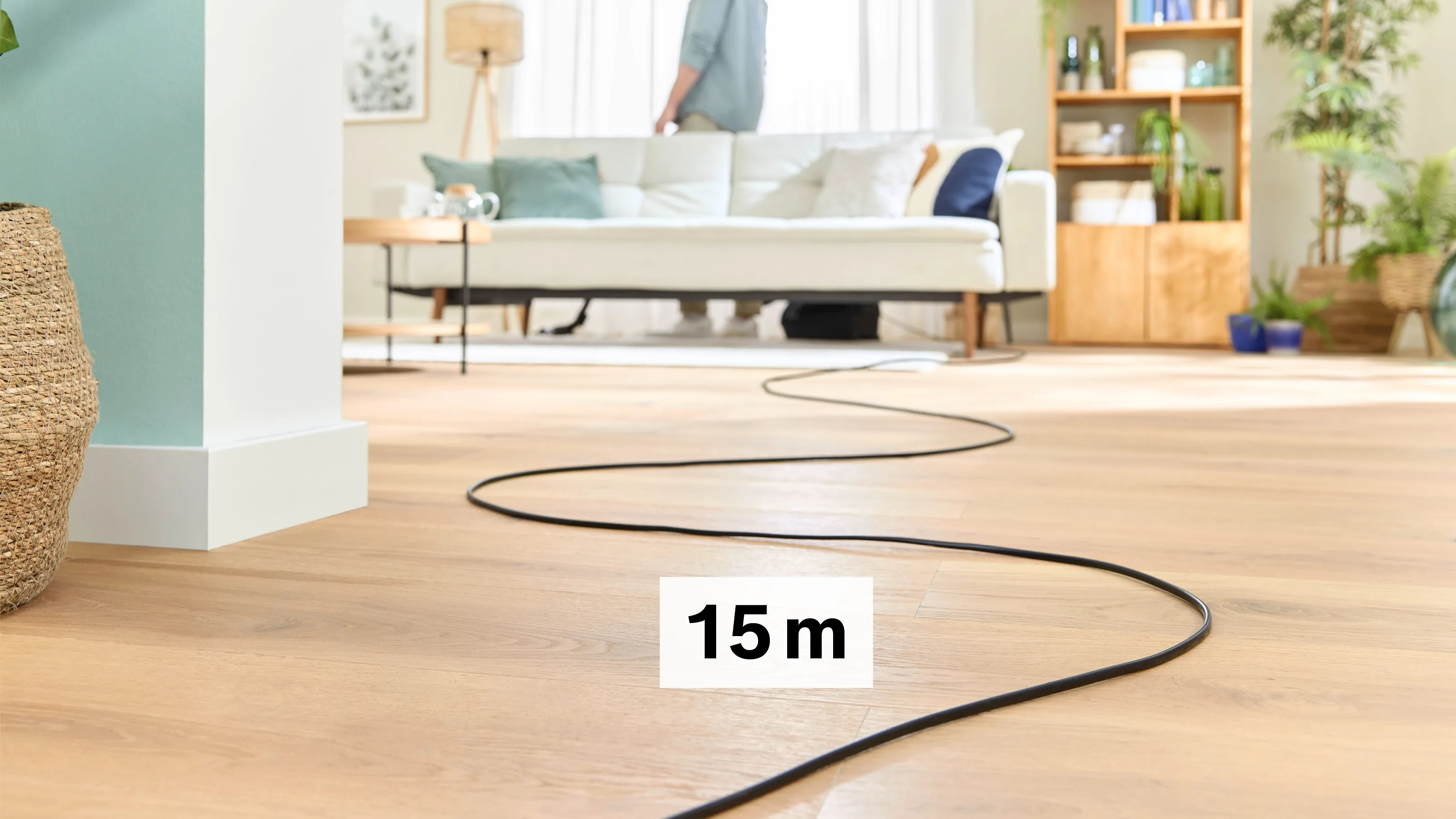 Un cable negro se extiende por un suelo de madera. Una persona está cerca de un sofá blanco.
