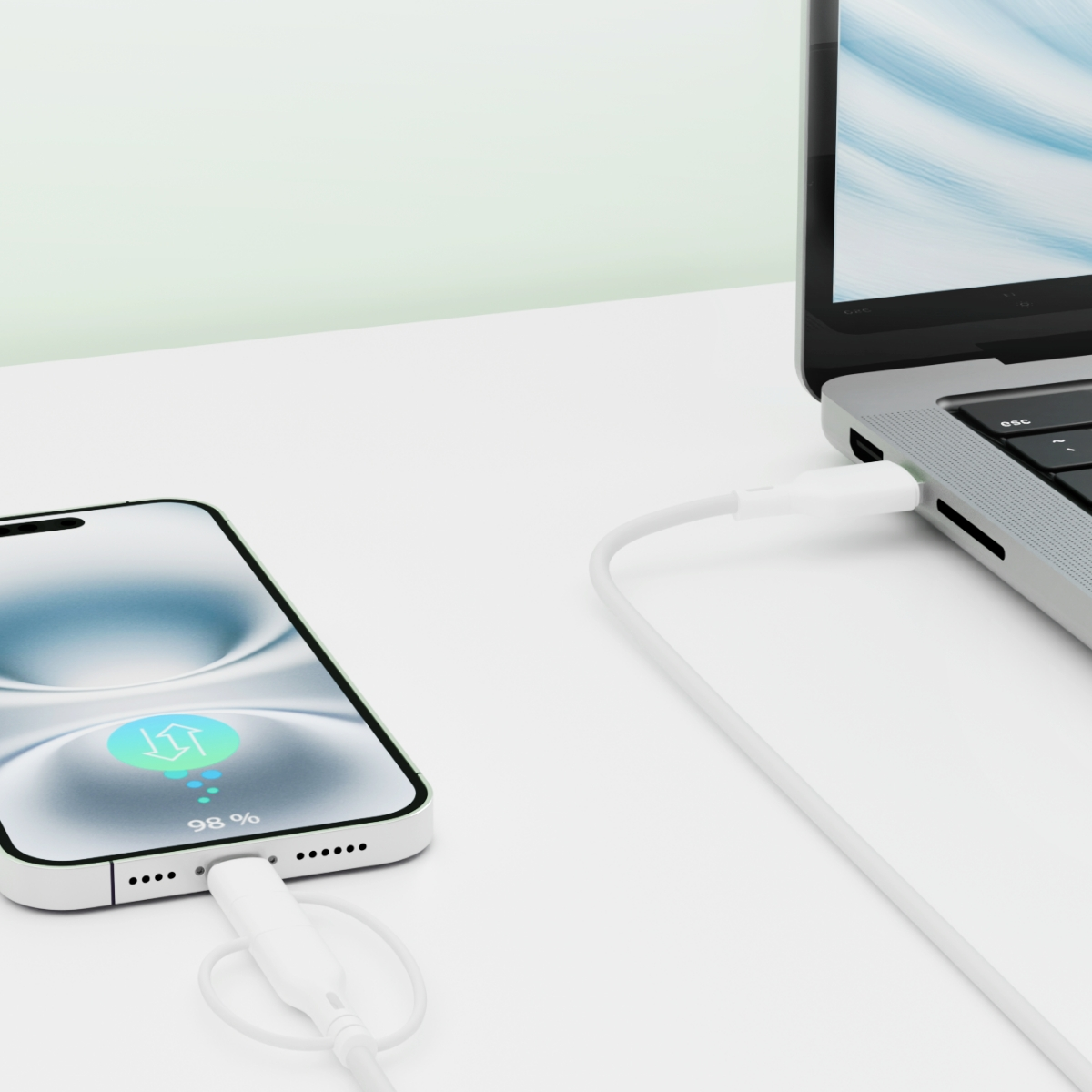 Telefon ładujący się obok laptopa. Biały kabel, telefon pokazuje ładowanie, białe biurko.