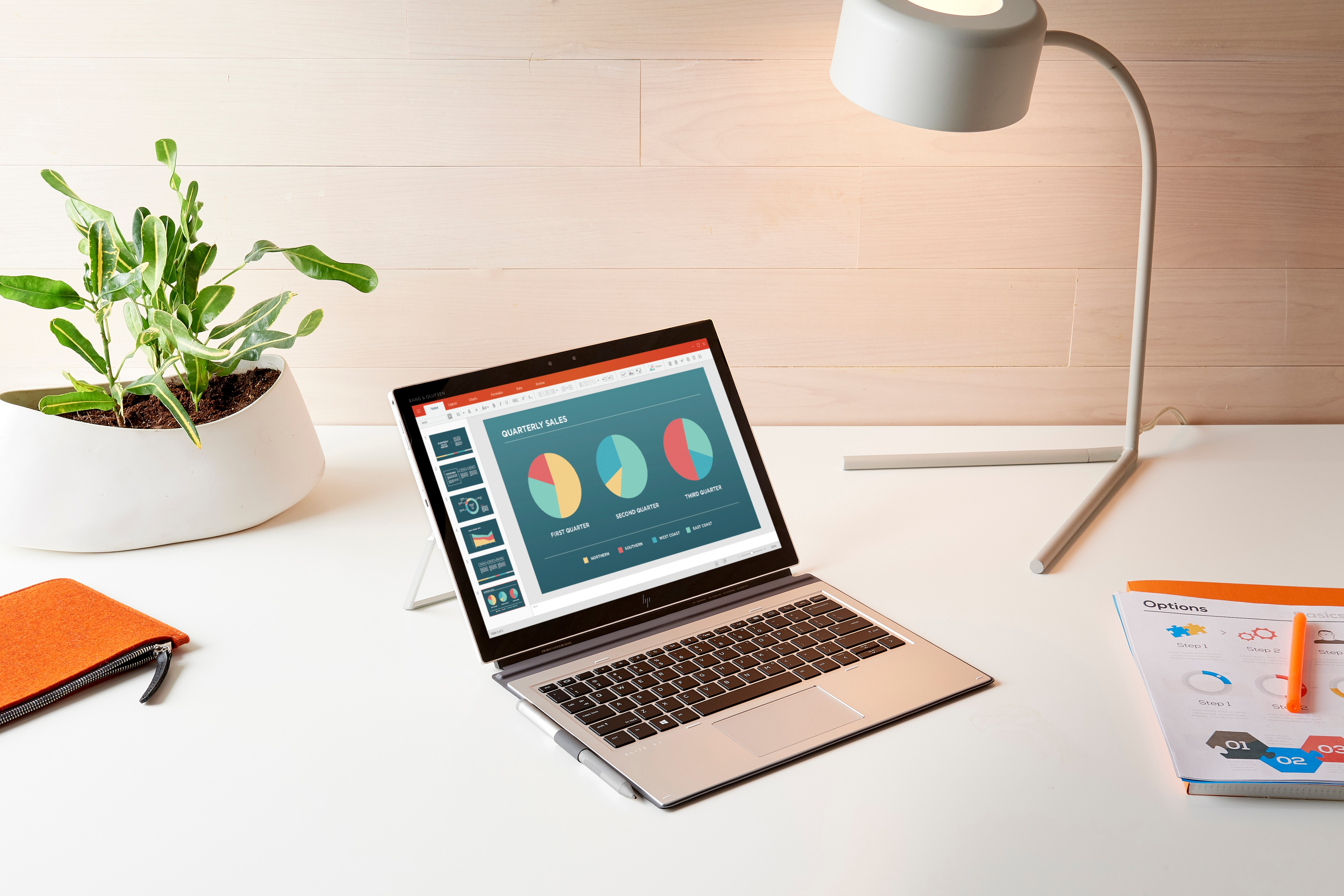 Laptop wyświetlający prezentację na białym biurku z lampą i rośliną.