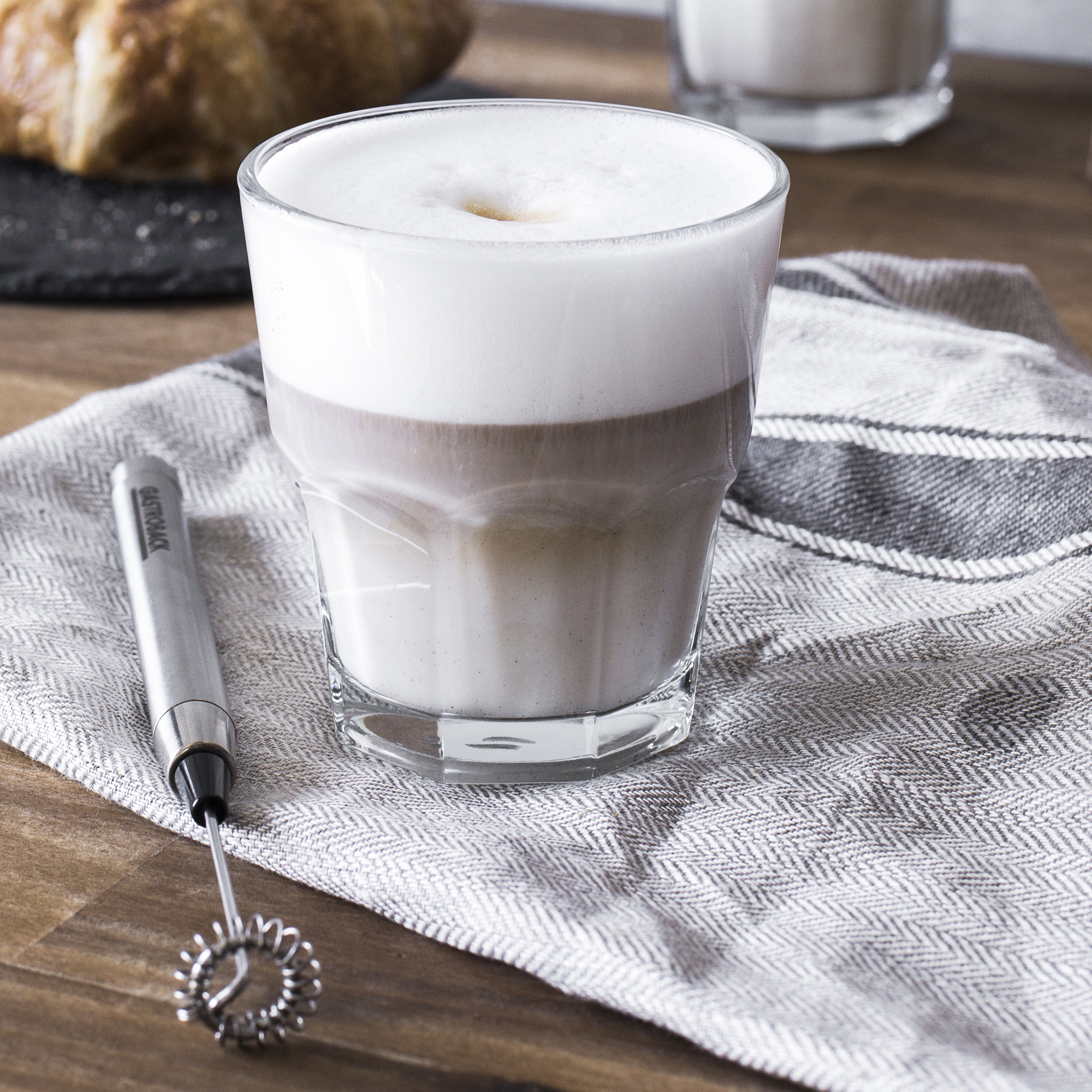 Un bicchiere di caffè a strati, con una parte superiore schiumosa e un montalatte e pane sul retro.