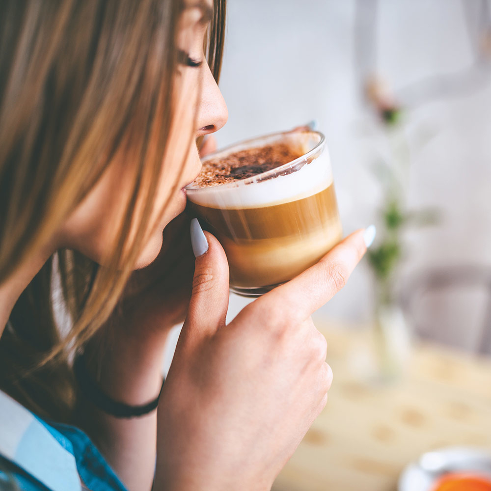 Donna che beve una bevanda al caffè a strati da un bicchiere trasparente, con uno sfondo sfocato.