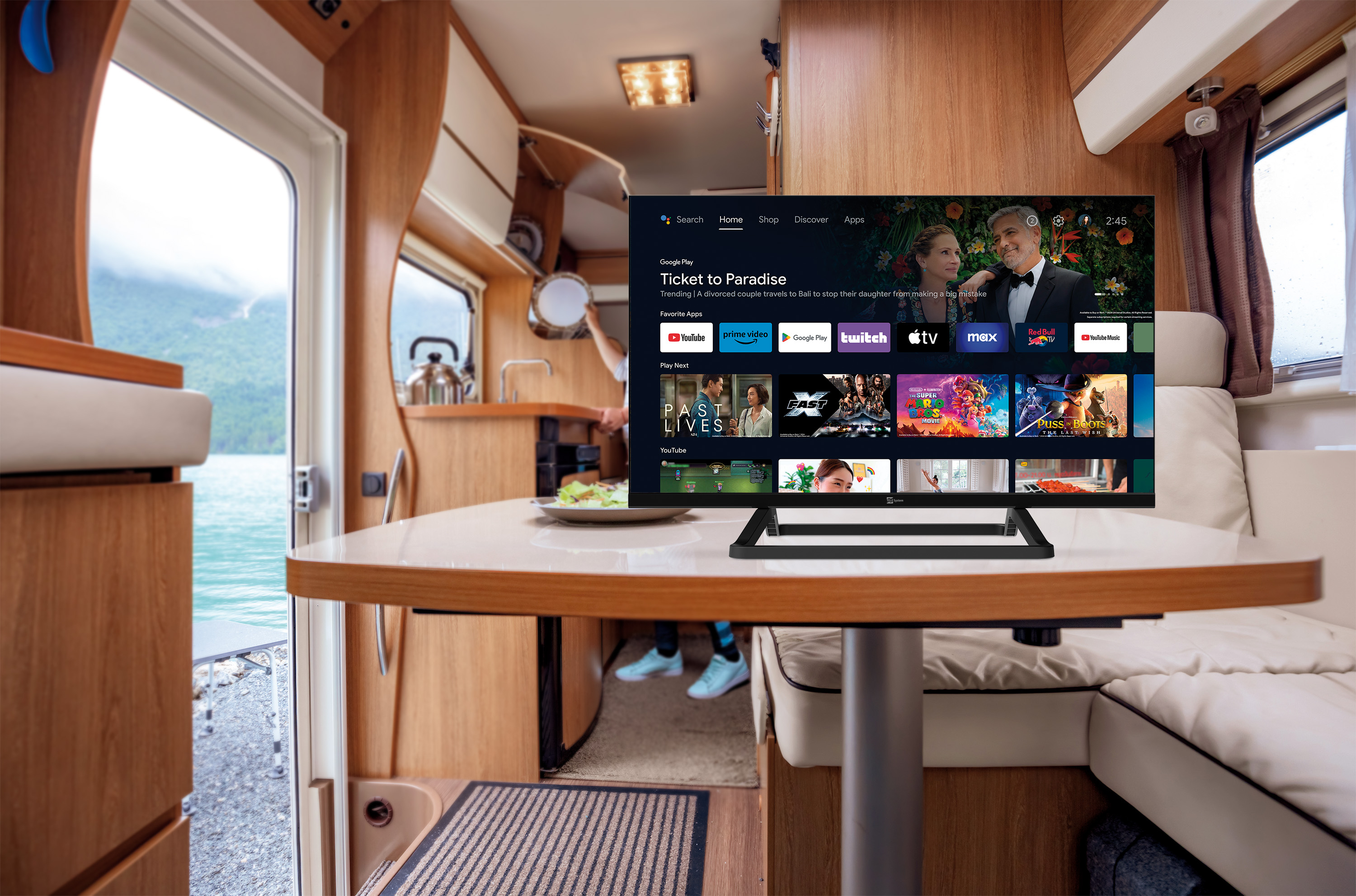 TV pokazuje 'Ticket to Paradise' w kamperze. Stół i drzwi na pierwszym planie.