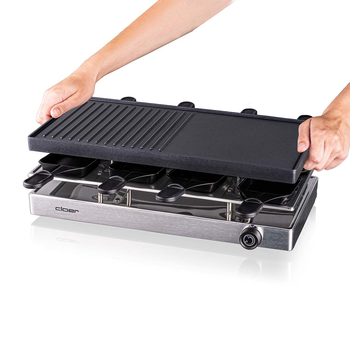 Mani che sollevano un coperchio nero per griglia da una griglia per raclette in acciaio inox, su sfondo bianco.