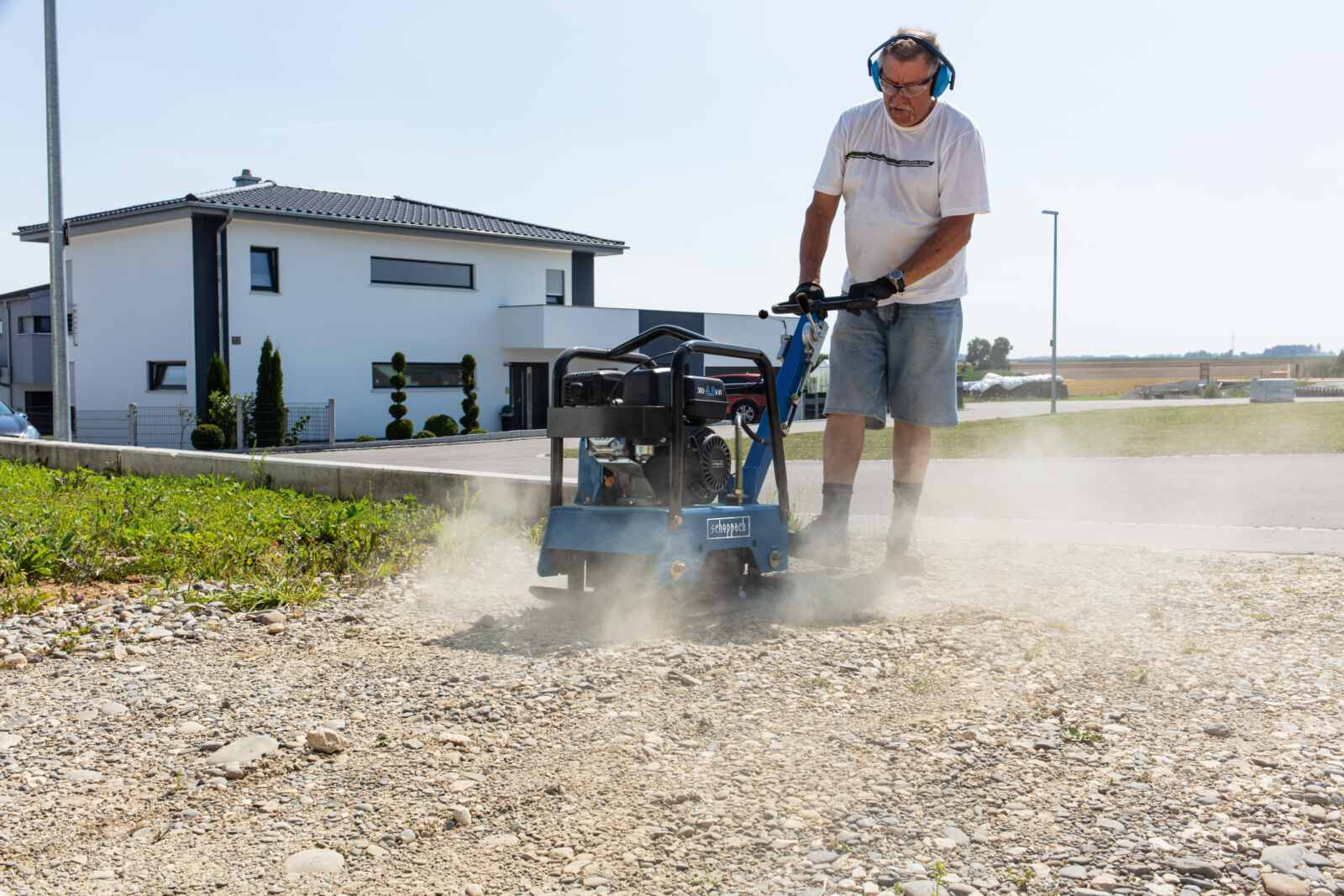 Een man die een blauwe Scheppach trilplaat gebruikt op grind, een gebouw op de achtergrond.