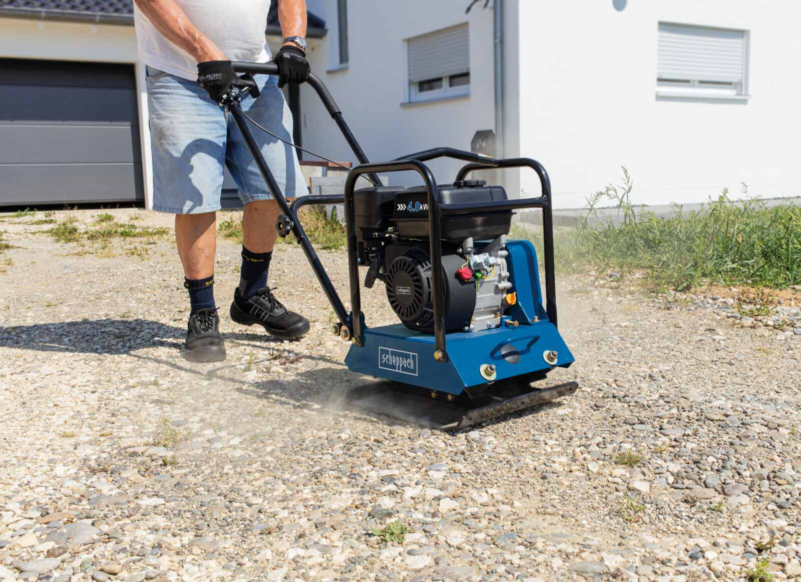 Man bedient blauwe en zwarte trilplaat op grind, buiten, zonnige dag.