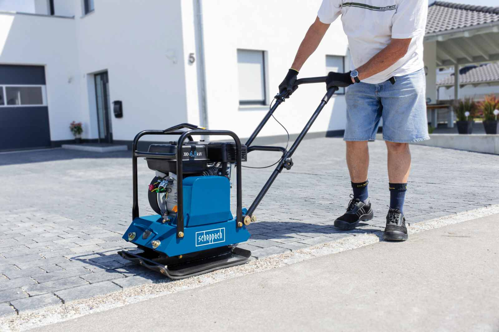 Man bedient een blauwe Scheppach verdichtingsmachine op een bestrate oprit.