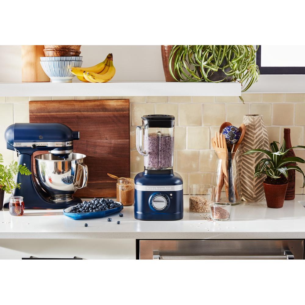 A kitchen scene with a mixer, blender, bananas, and other items on a white countertop. A vase and plants are present.