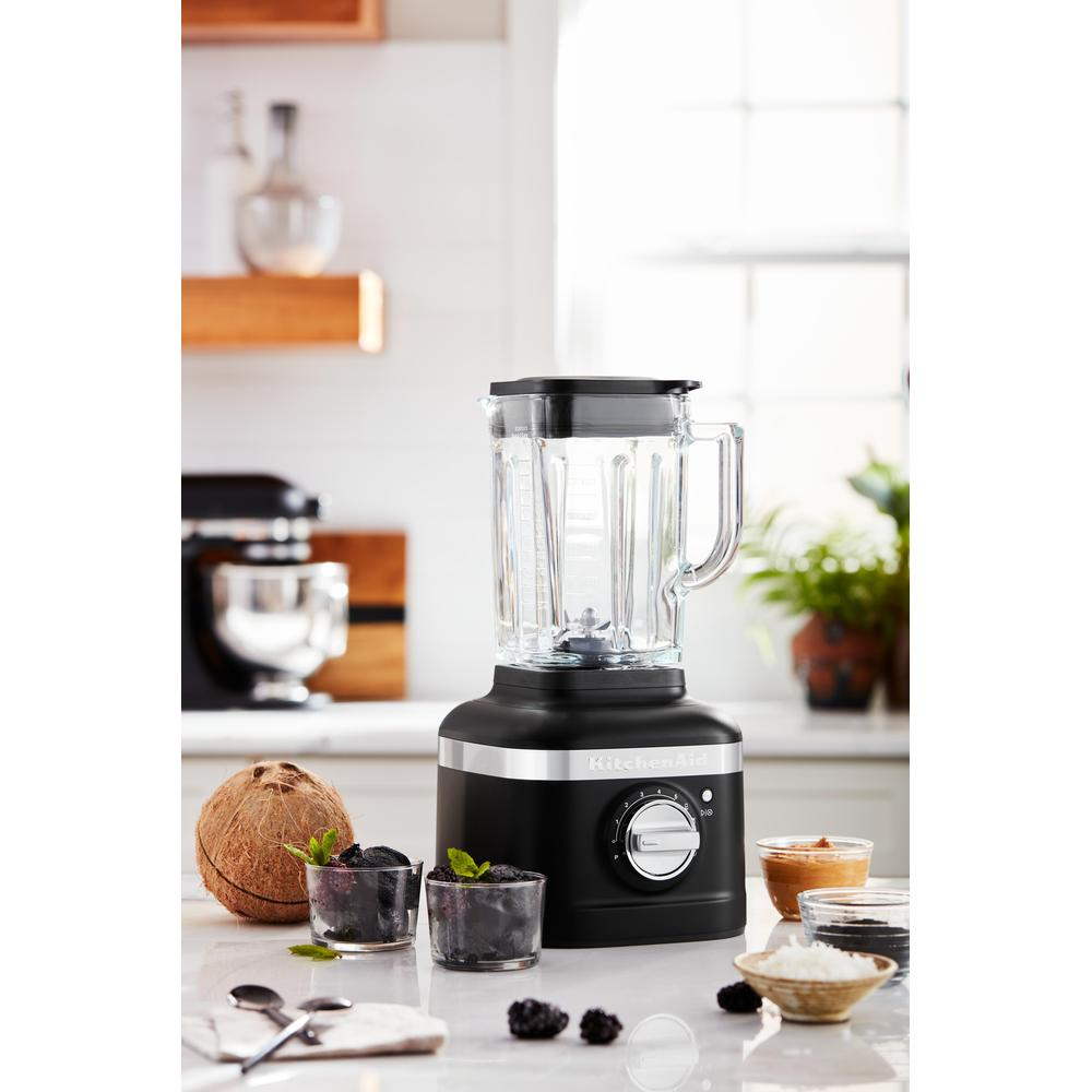 Schwarzer Mixer mit Glasbehälter auf weißer Oberfläche, mit Zutaten in der Nähe.