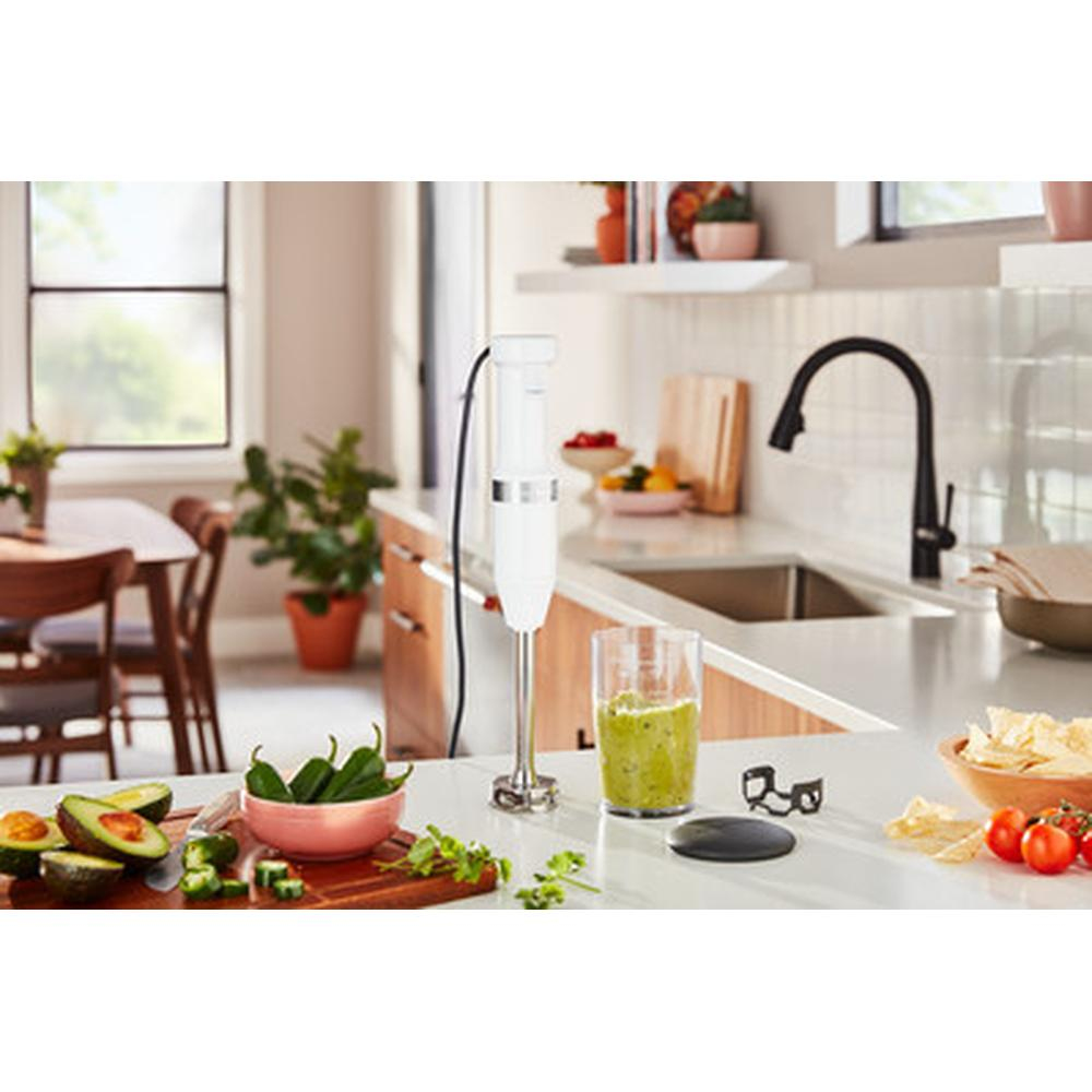 Scena di cucina con frullatore a immersione bianco, bicchiere con liquido verde e avocado.