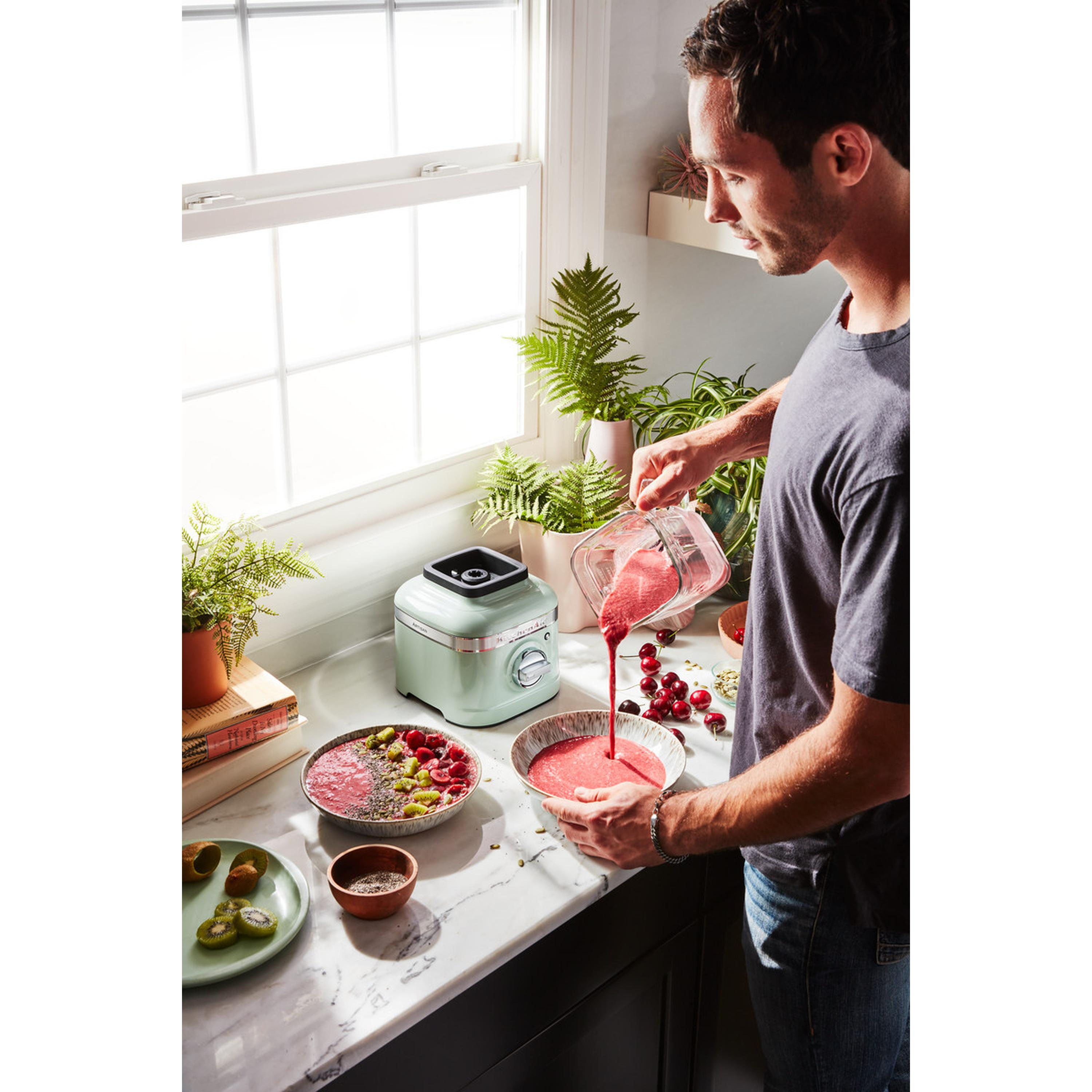 Man pouring pink smoothie into a bowl, next to a blender, kiwis and berries.
