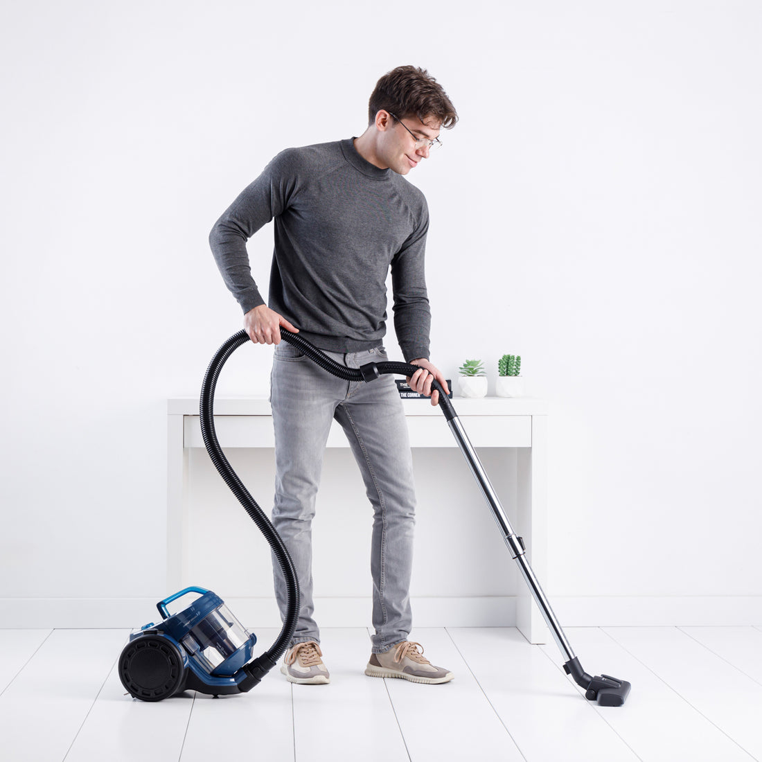 Mężczyzna odkurza białą podłogę niebieskim odkurzaczem. Ma okulary.