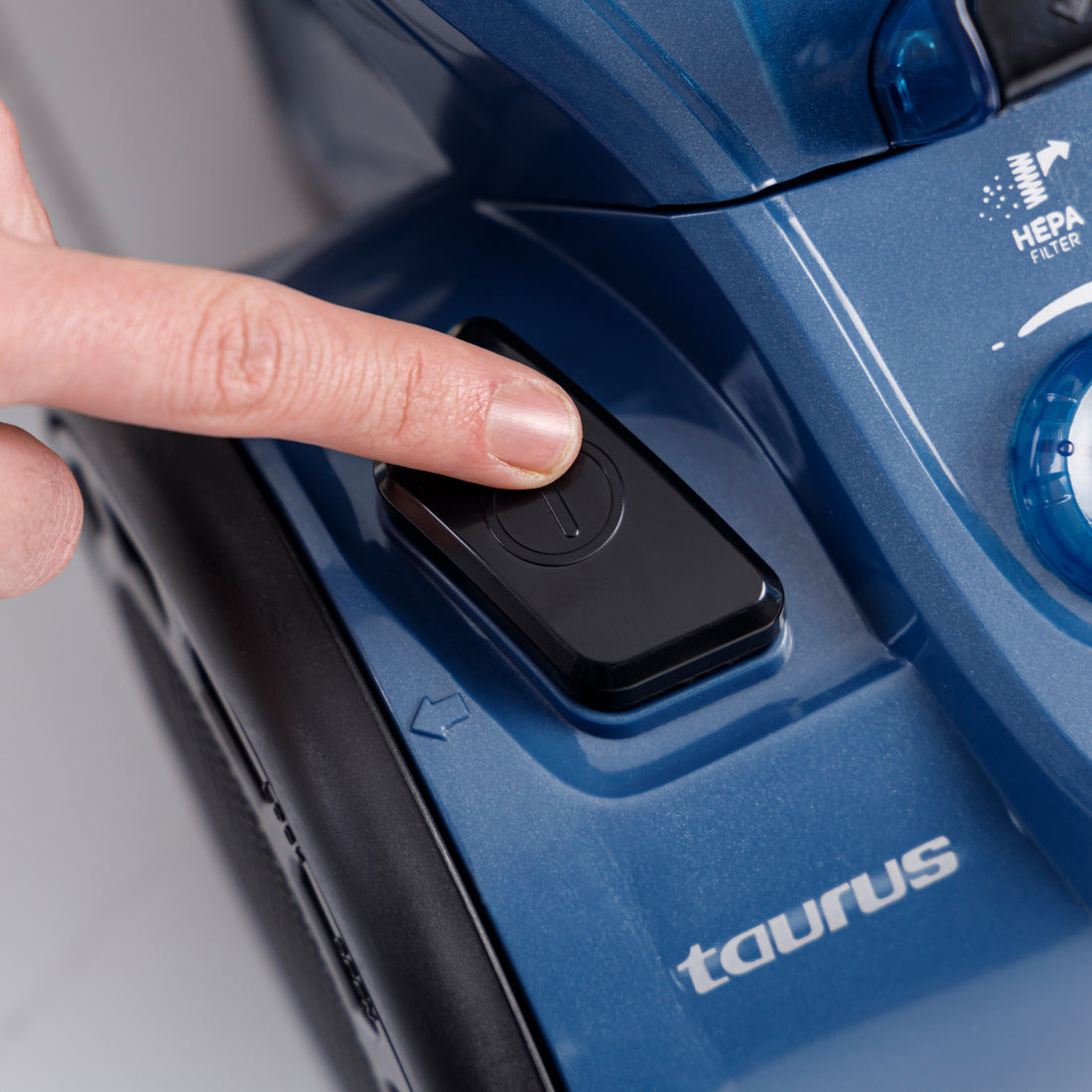 Ręka naciska czarny przycisk zasilania na niebieskim odkurzaczu. Logo to Taurus.