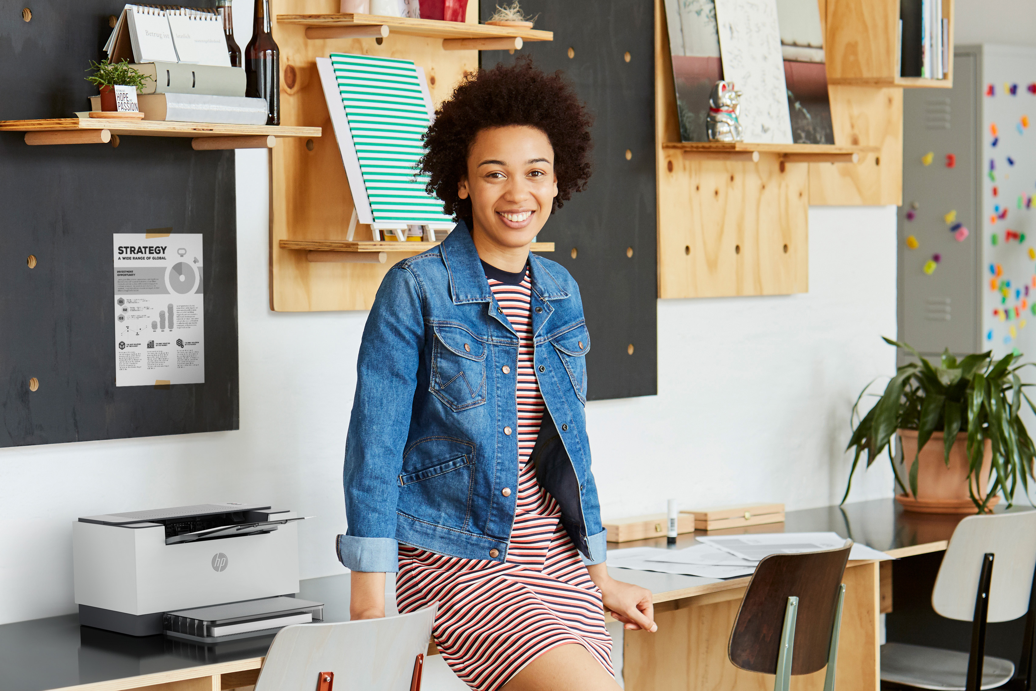 Kobieta w dżinsowej kurtce stojąca przy biurku uśmiecha się. Drukarka i dokumenty na biurku.