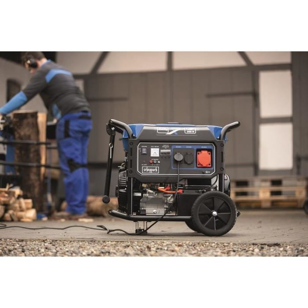 Een zwarte en blauwe generator op wielen. Een man werkt op de achtergrond bij een stapel hout.