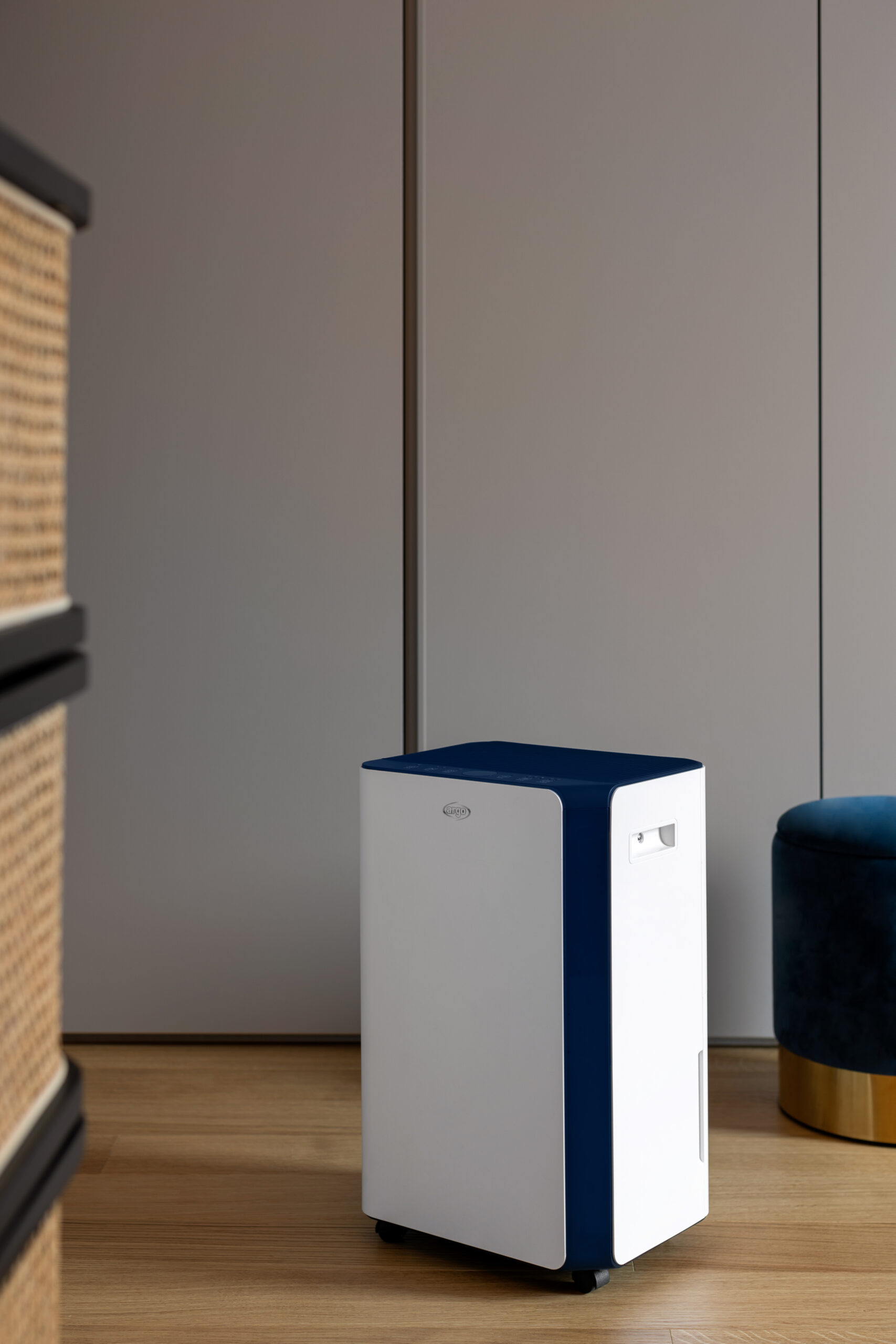Een witte en blauwe luchtontvochtiger staat op een houten vloer. Een grijze muur op de achtergrond, een blauwe kruk.