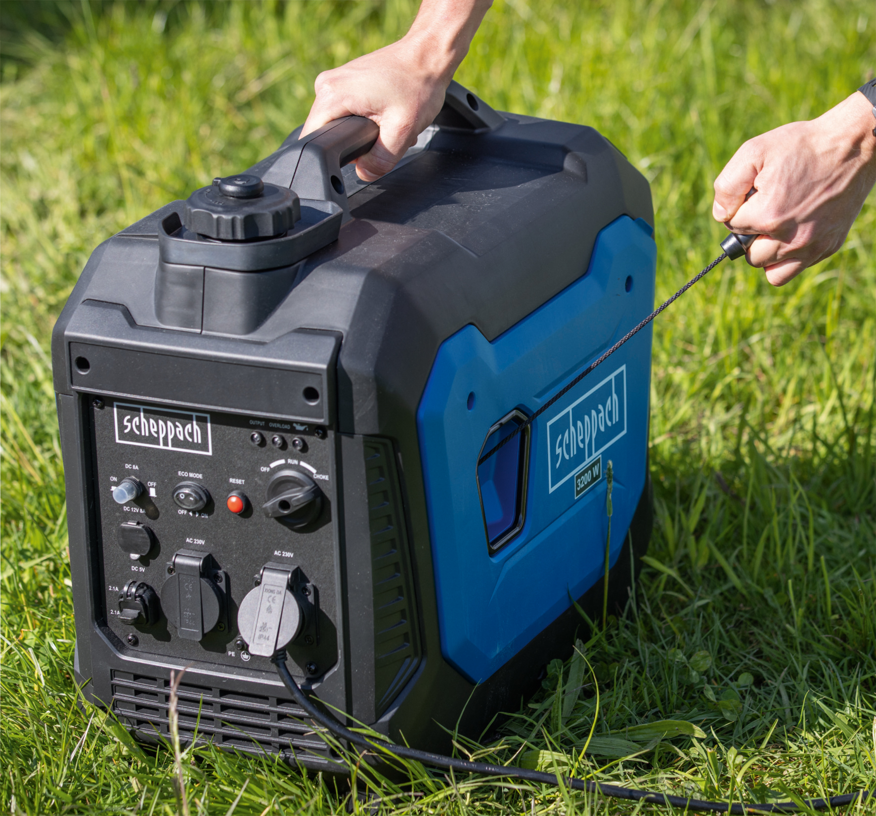 Een blauw-zwarte generator op gras. Een hand trekt aan het startsnoer.