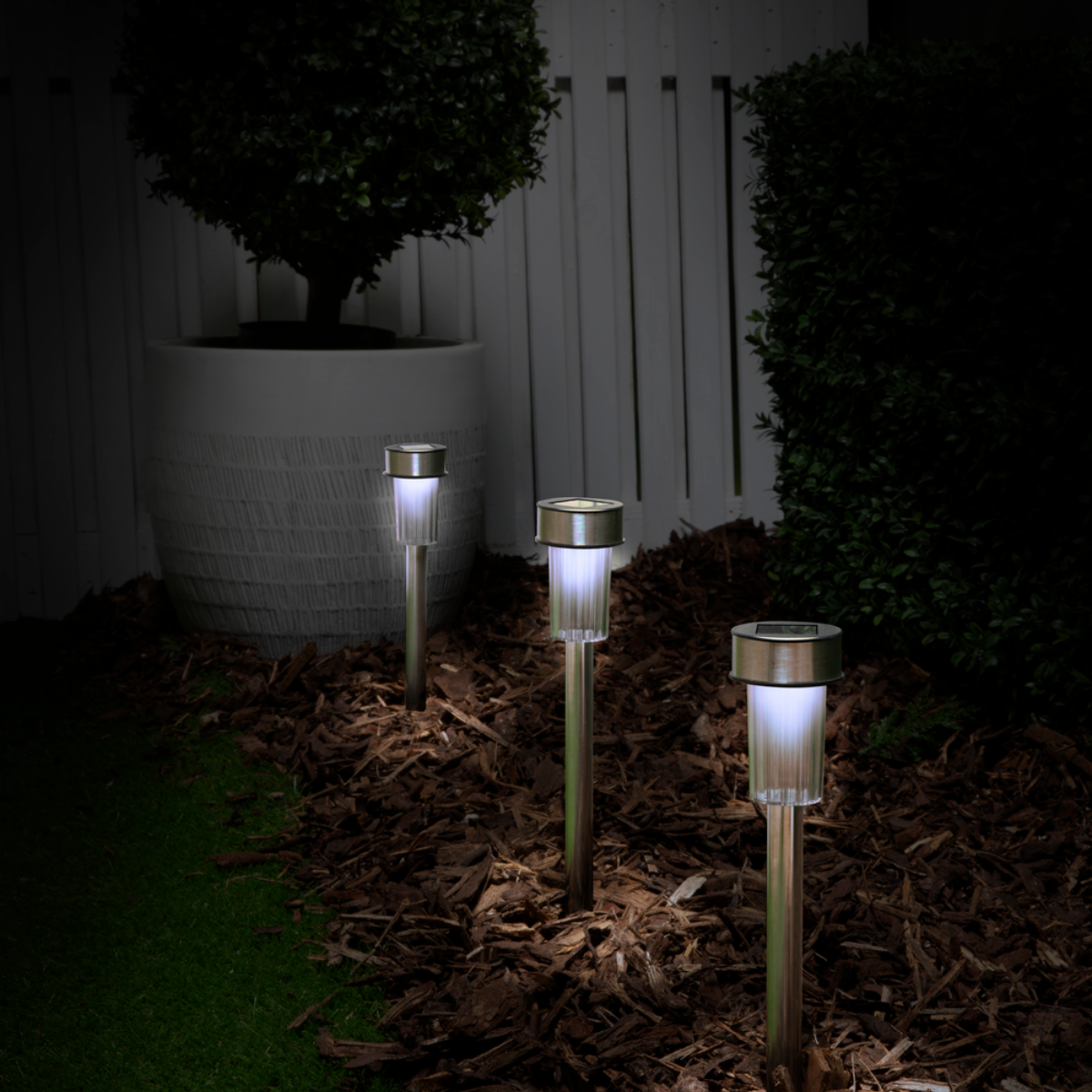 Drie verlichte zonne-tuinlampen in een donkere tuin, met een grote bloempot en hek op de achtergrond.