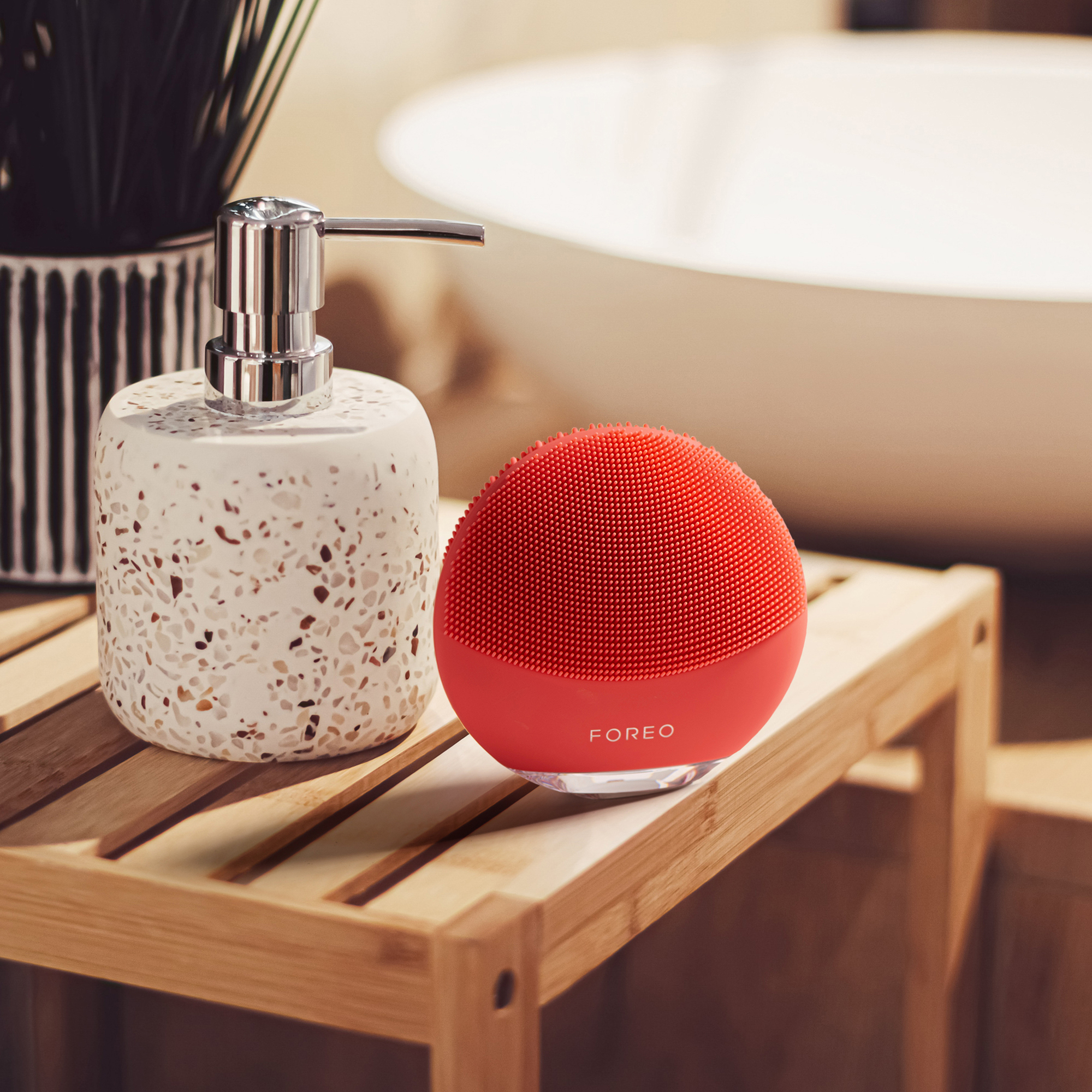 Red facial cleansing brush and soap dispenser on a wooden shelf. White background.