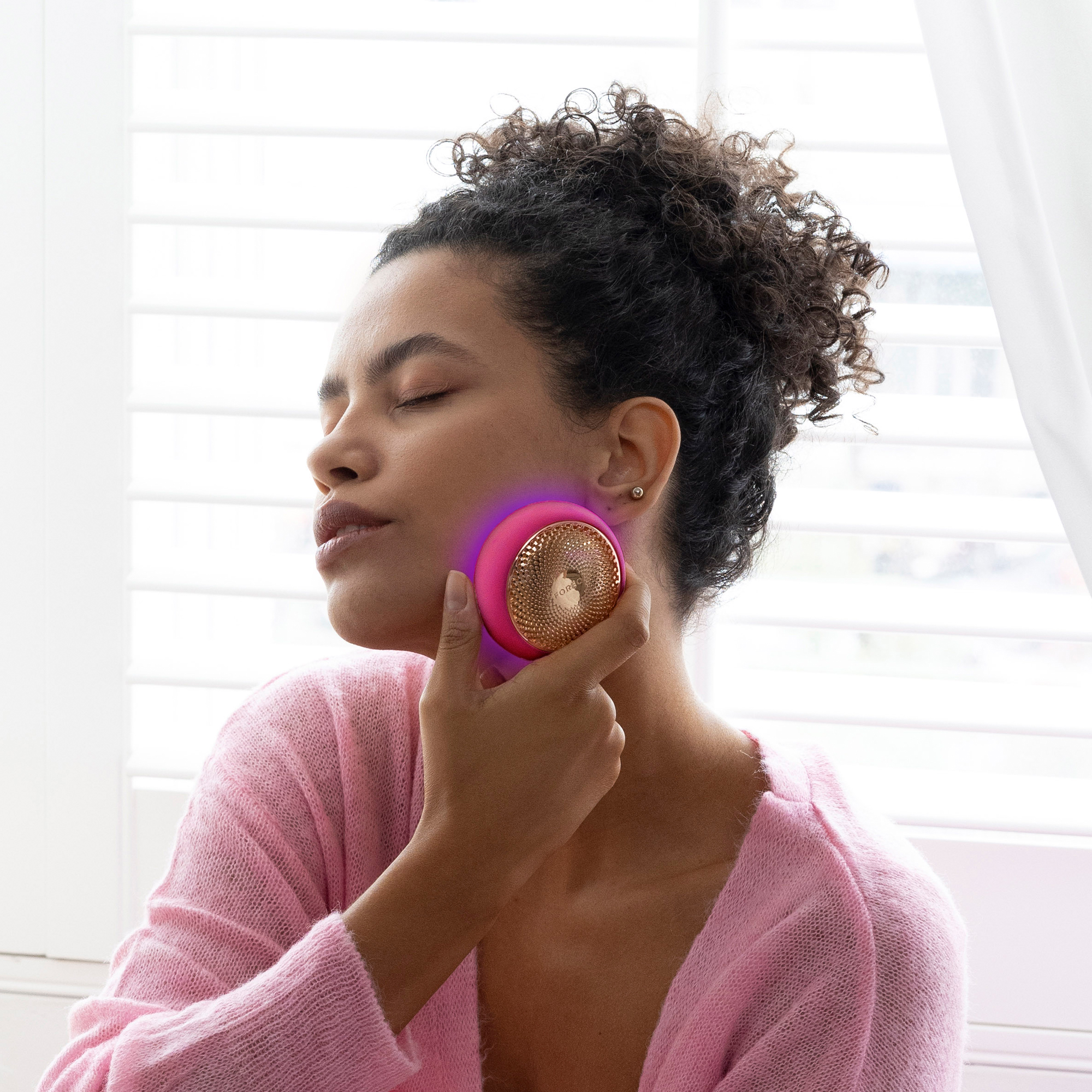 Woman holding a pink skincare device on her face. Eyes closed. Window blinds in the background.