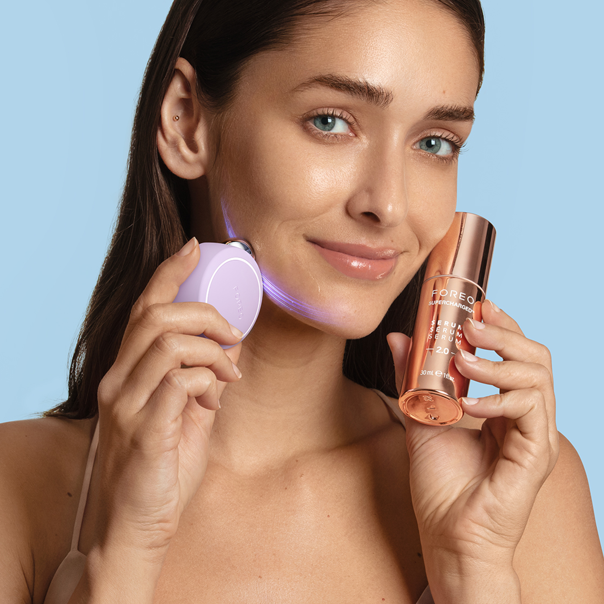 Une femme tient un appareil facial lilas et une bouteille de sérum or rose sur fond bleu.