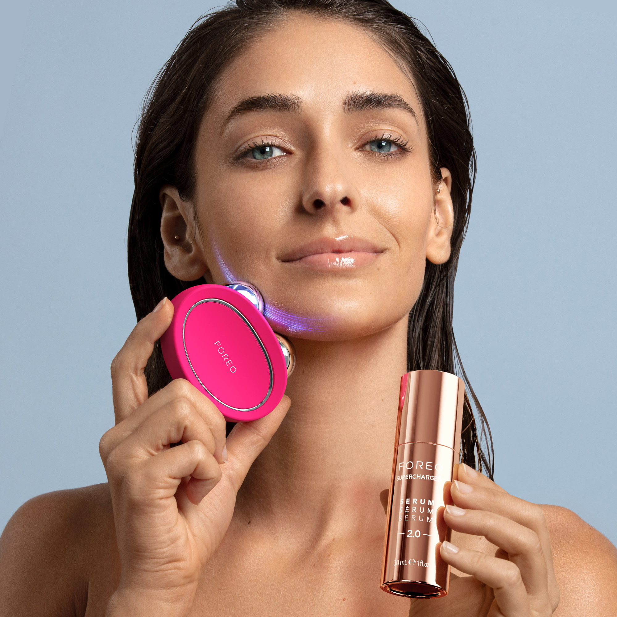 Woman holds a pink facial device and rose gold serum bottle on a blue background.