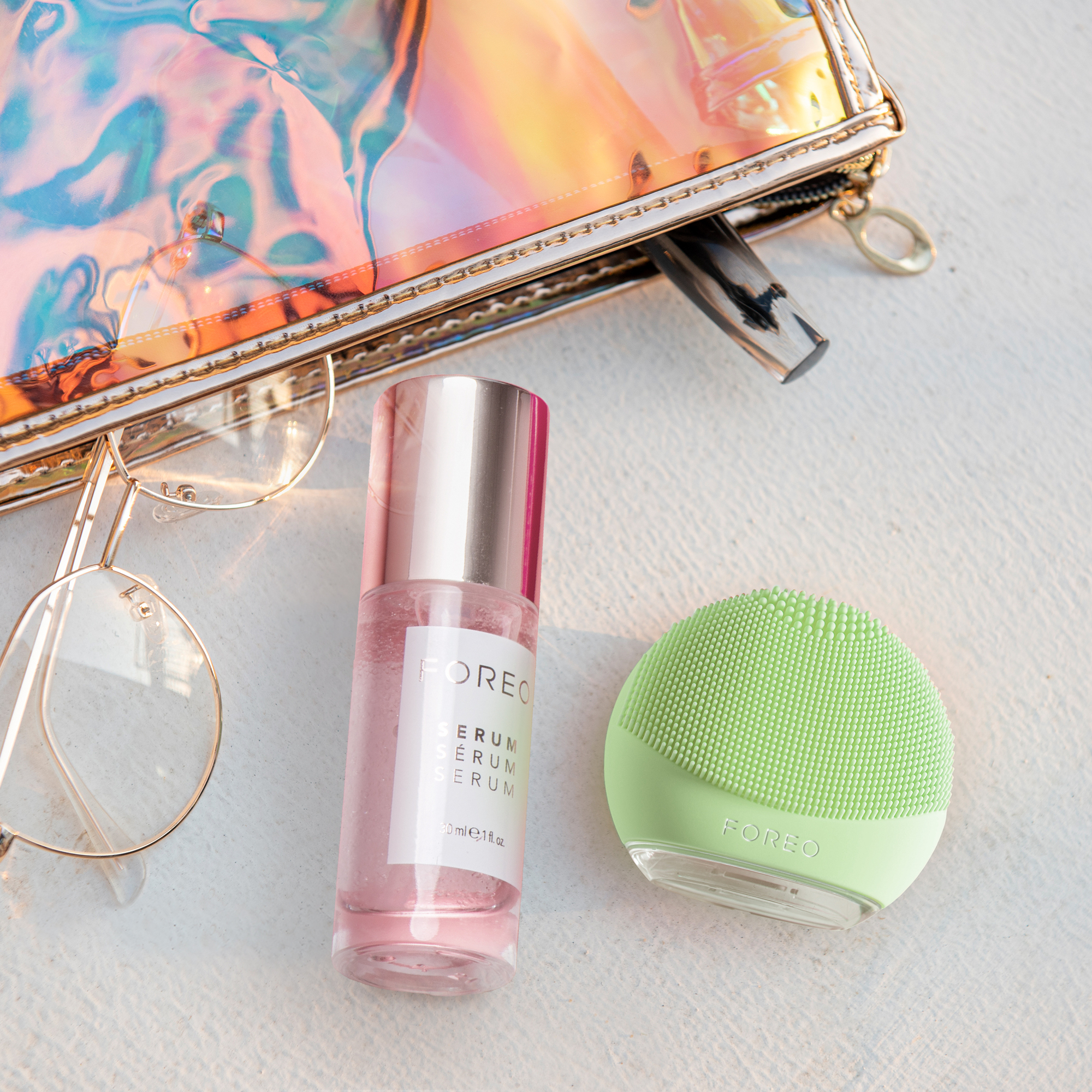 Foreo Luna cihazı, serum şişesi ve gözlük, şeffaf bir çantayla beyaz yüzeyde.