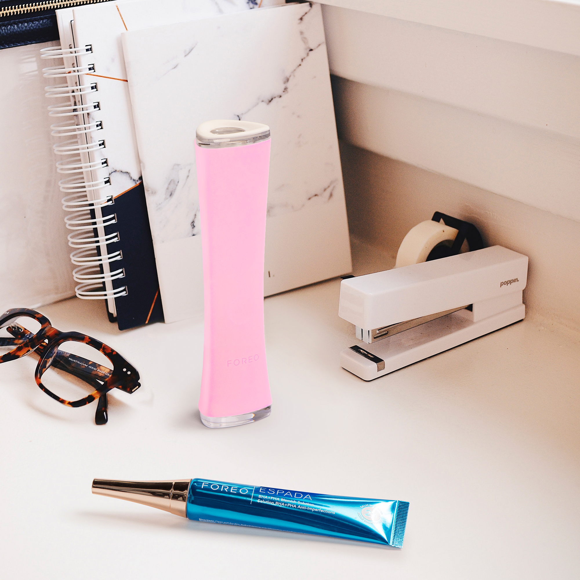 Desk scene with a pink Foreo Espada device, glasses, stapler, and notebook.