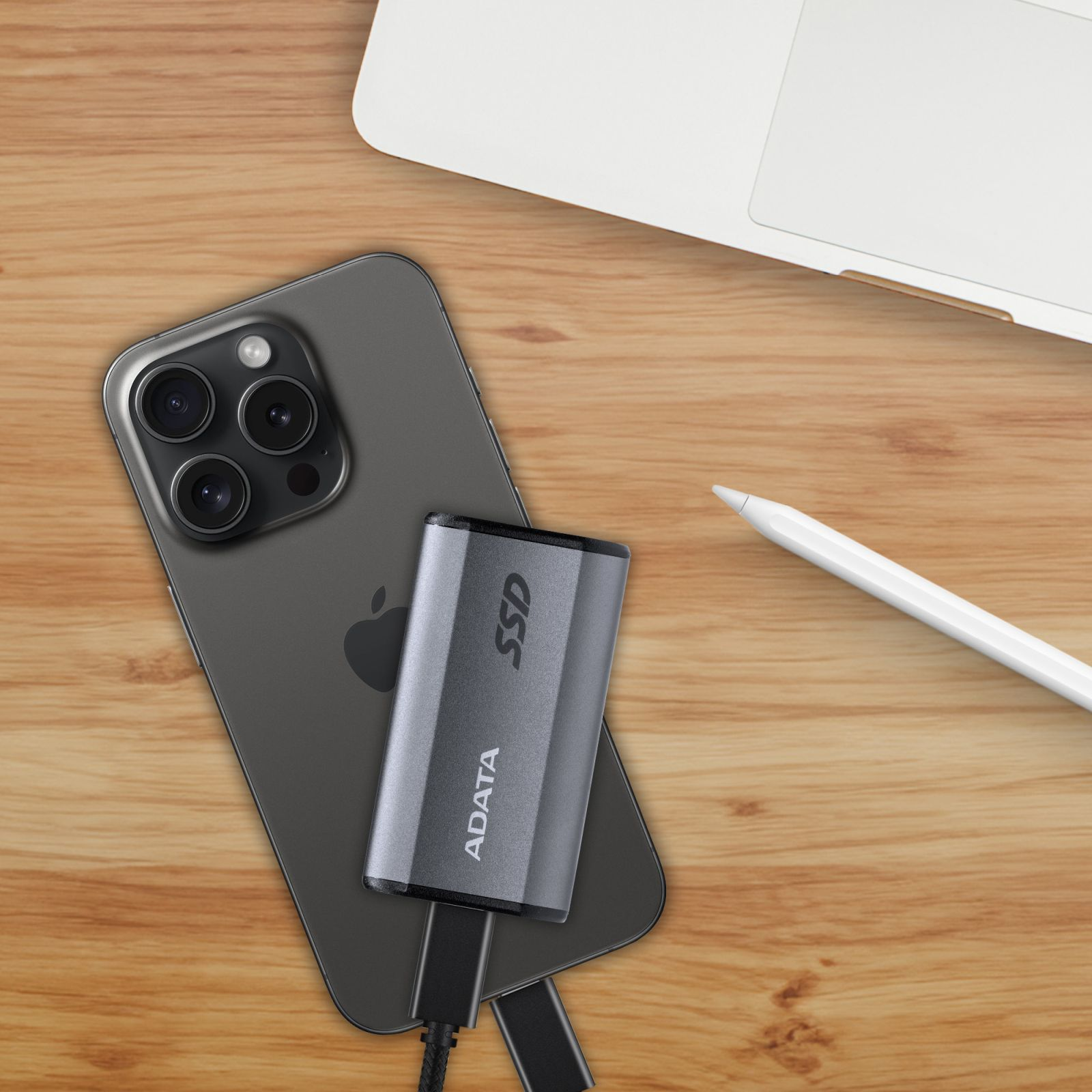 Szary dysk SSD jest podłączony do czarnego iPhone'a na drewnianym stole, z białym rysikiem i laptopem.