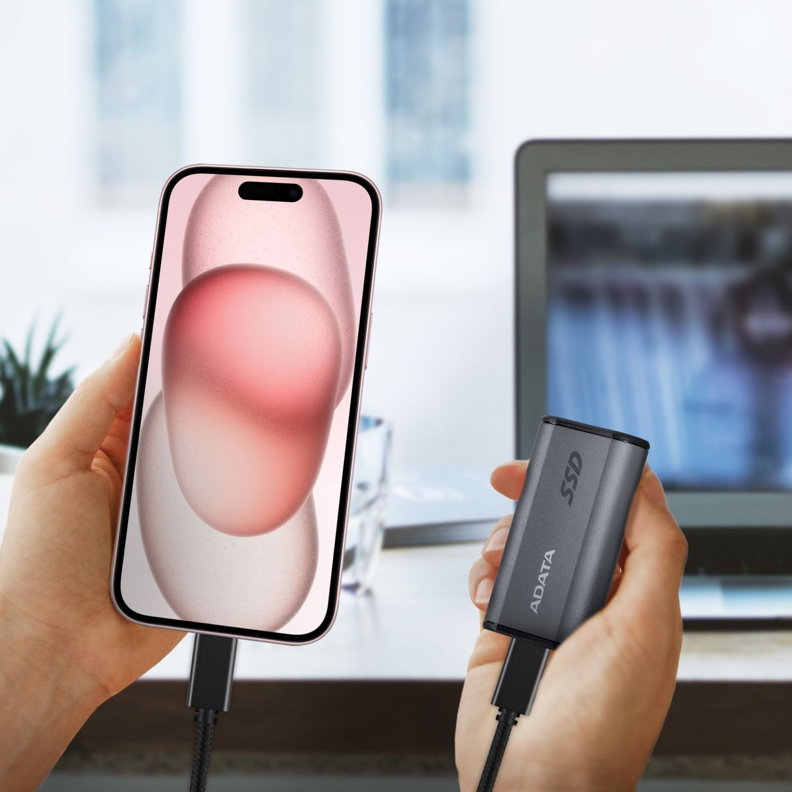 Dłonie trzymające iPhone'a podłączonego do szarego dysku SSD, z rozmytym laptopem w tle.