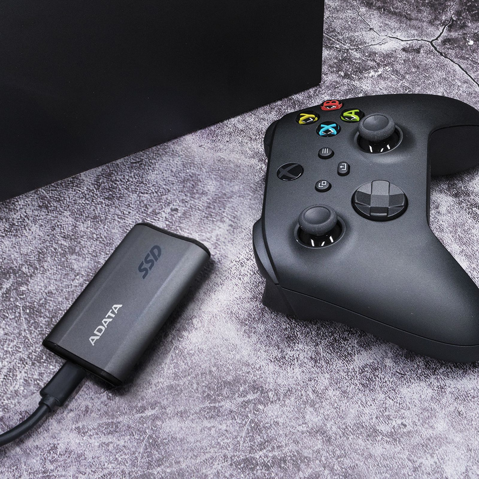 Szary dysk SSD podłączony do czarnego kontrolera Xbox, leżący na teksturowanej powierzchni.