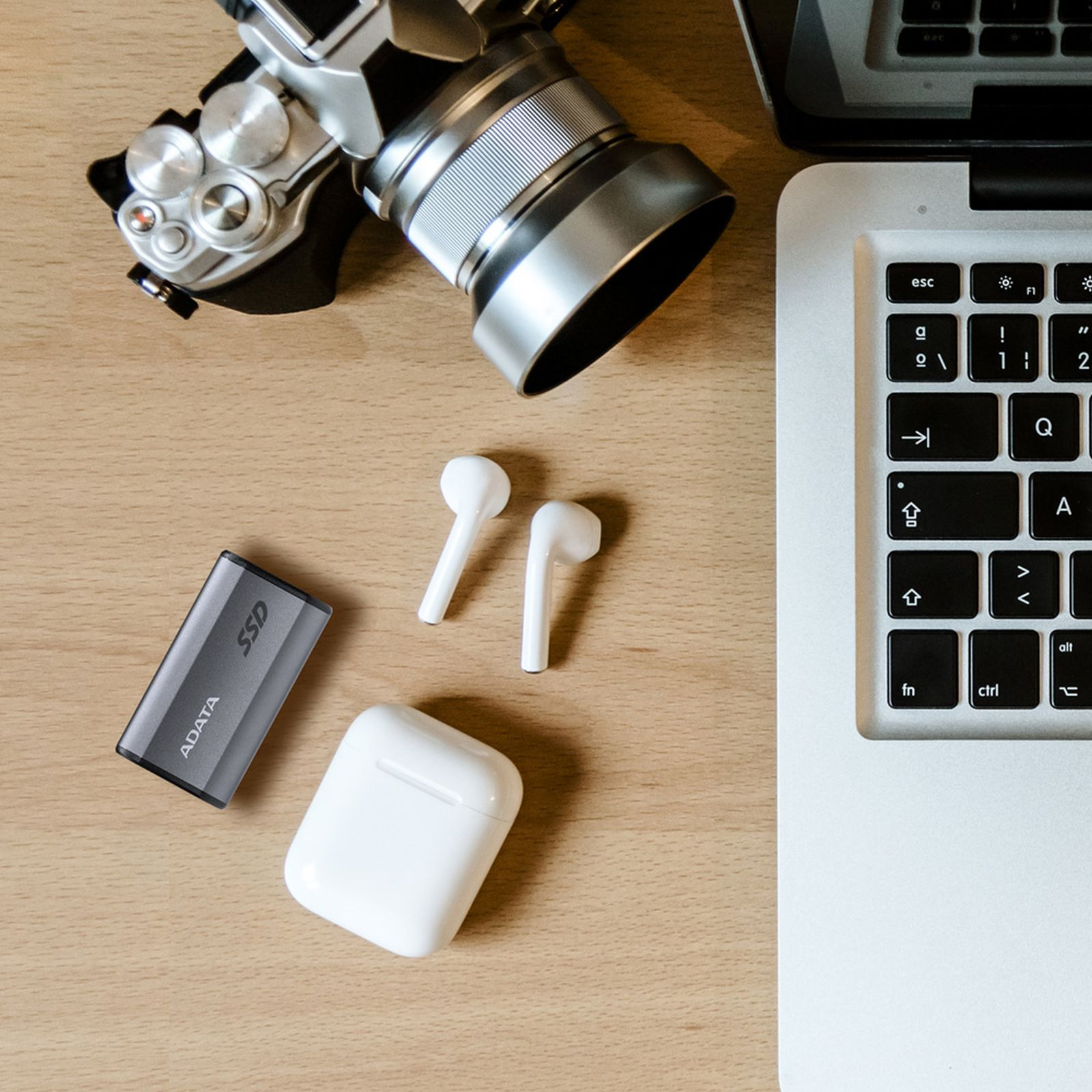 Widok z góry na biurko z aparatem, dyskiem SSD, słuchawkami AirPods, etui i laptopem.