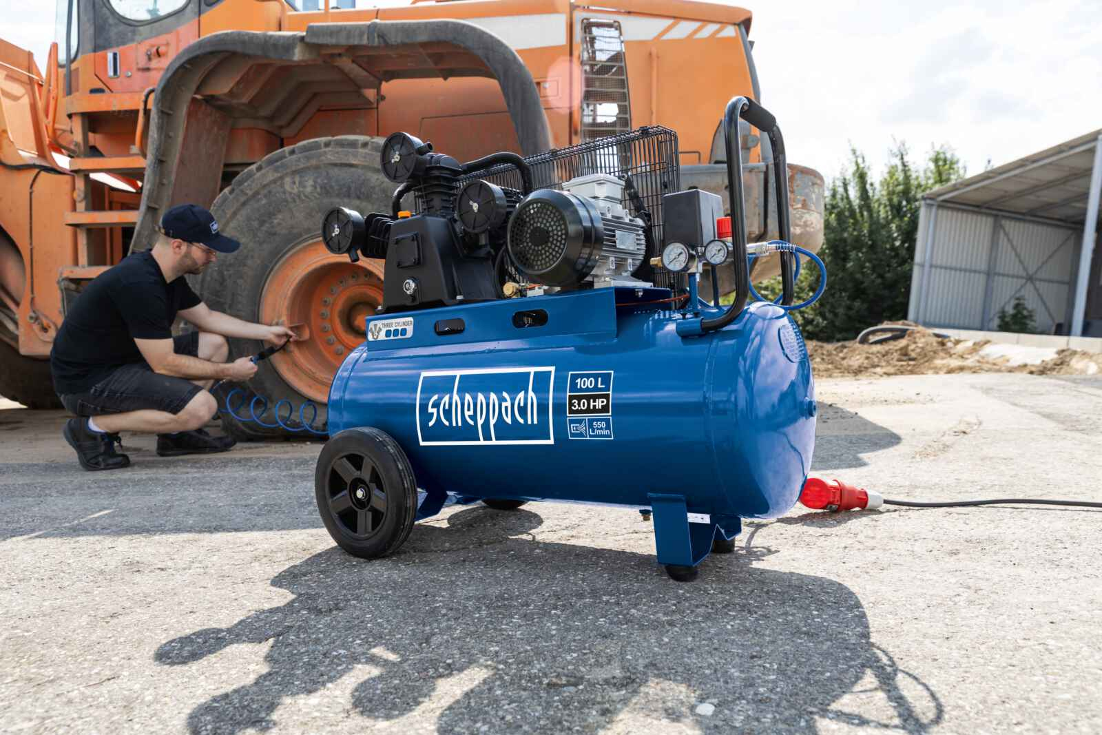 Een blauwe Scheppach luchtcompressor op beton, persoon werkt aan een band.