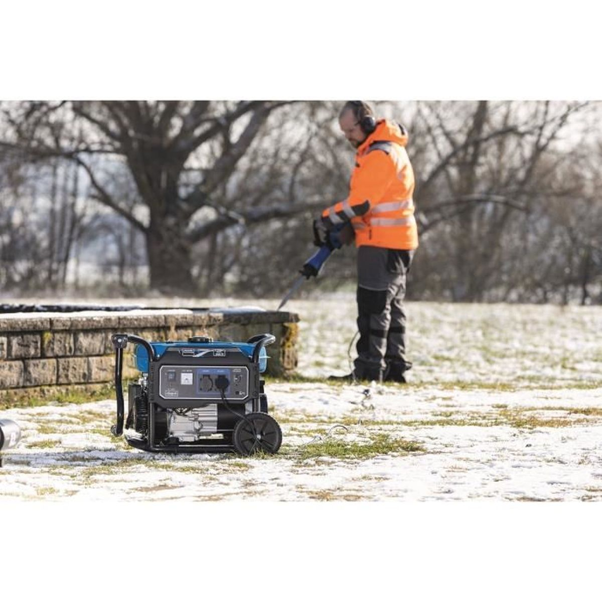 A person in orange workwear uses a tool near a generator in a snow-covered area.